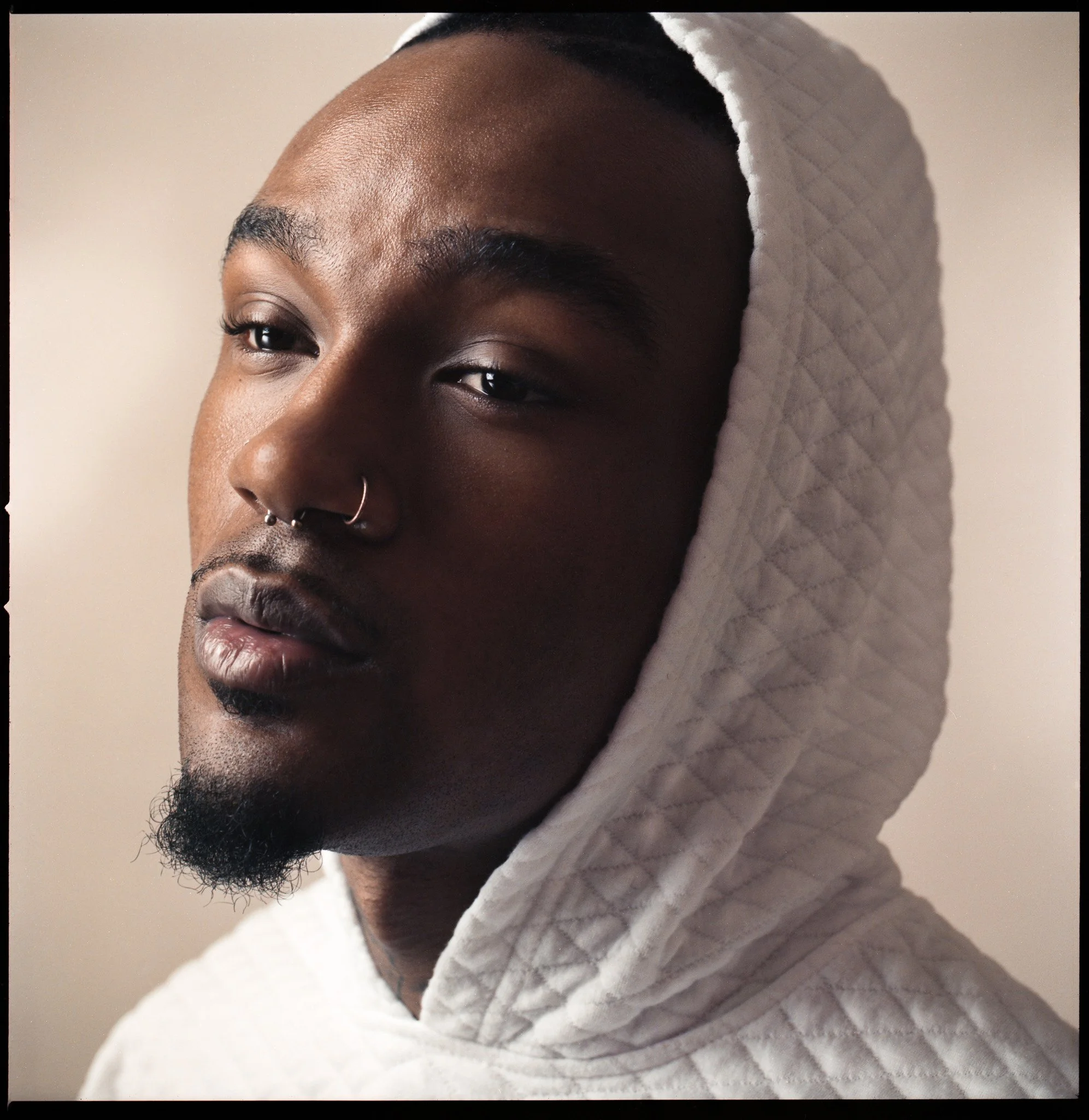Close-up of a young Black man with a nose ring and styled facial hair, wearing a quilted white hoodie with the hood up, against a neutral background.