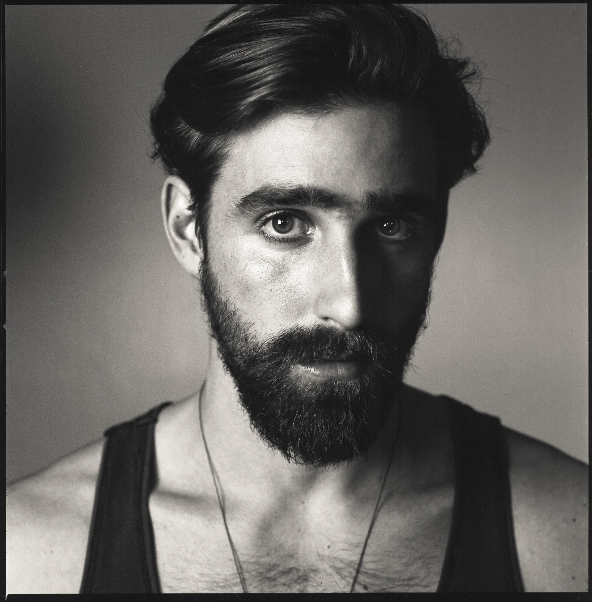 Black and white portrait of a young man with dark hair, a beard, wearing a sleeveless shirt and a necklace, looking directly at the camera.