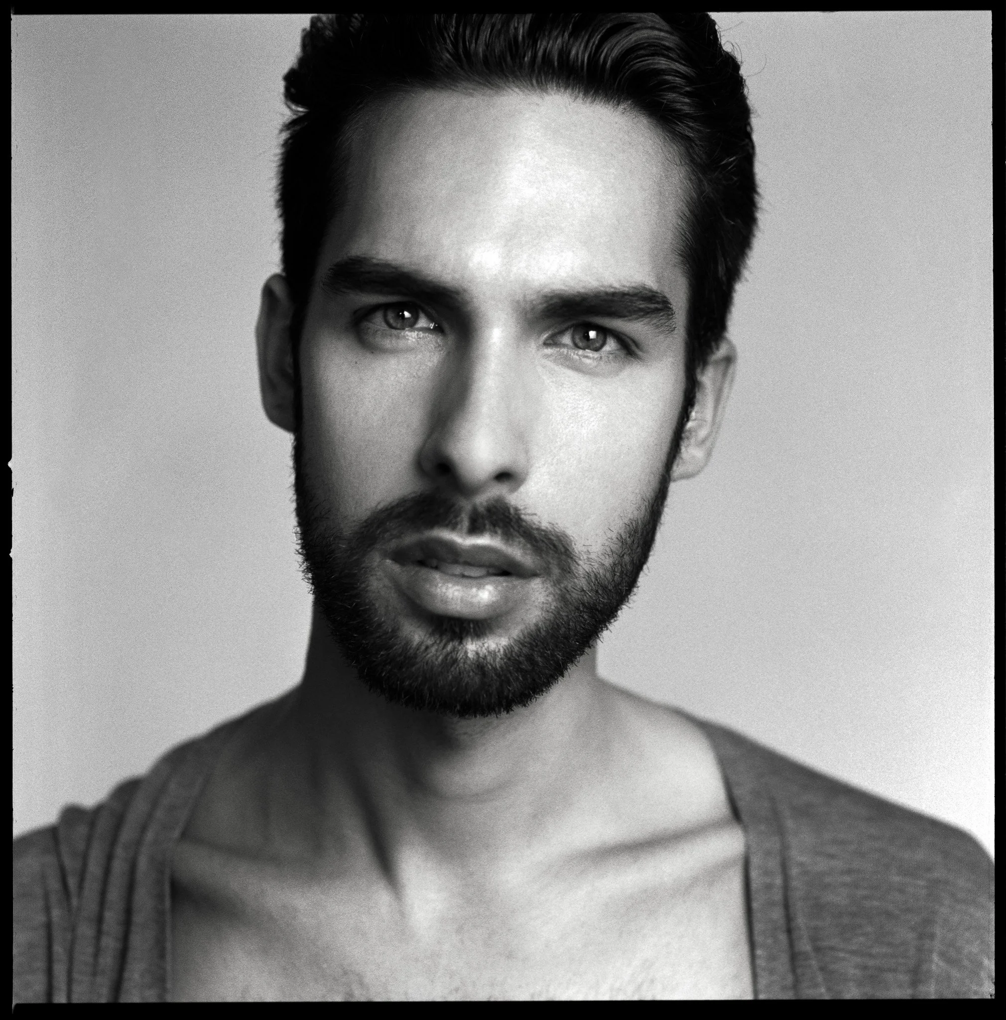 Black and white portrait of a young man with facial hair, wearing a casual shirt, looking directly at the camera with a neutral expression.