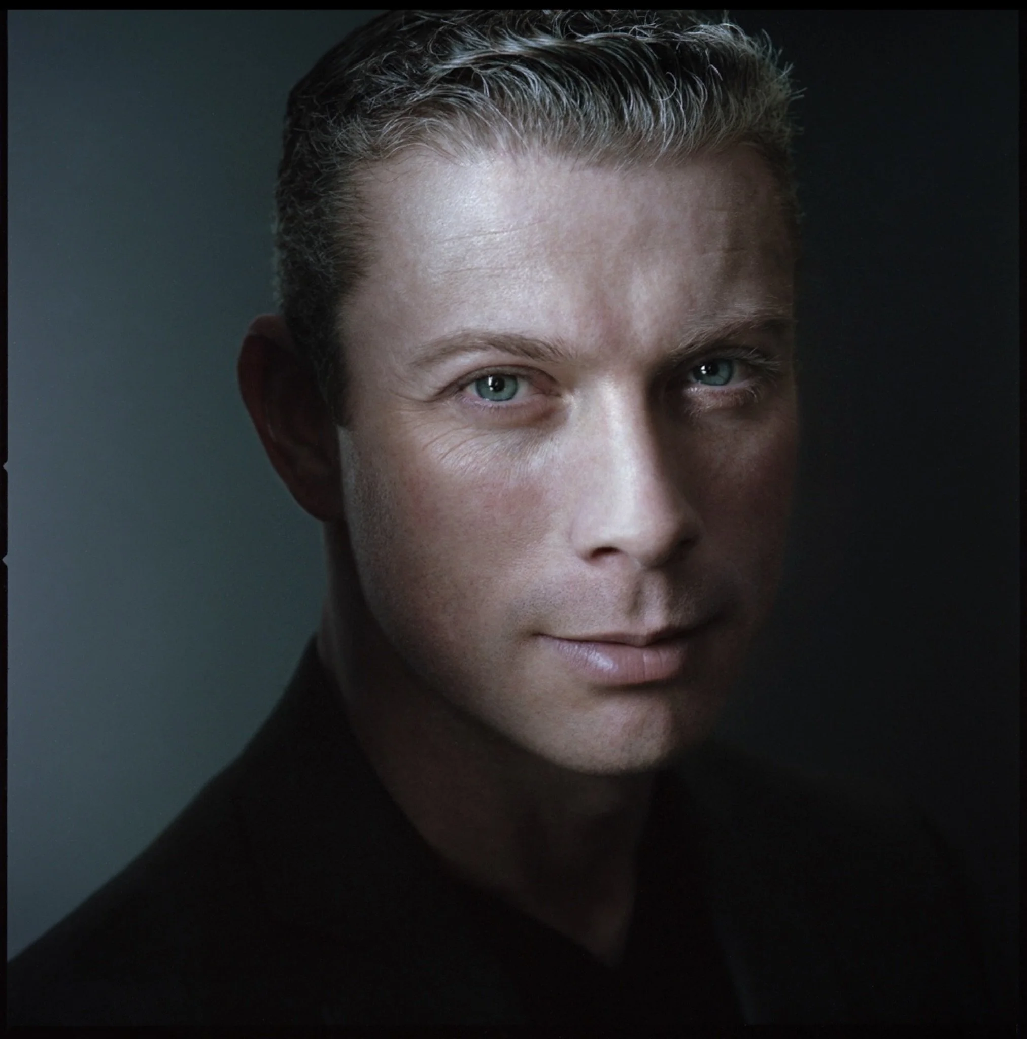 Close-up portrait of a man with blue eyes and short, slicked-back hair, wearing a black shirt, against a dark background.