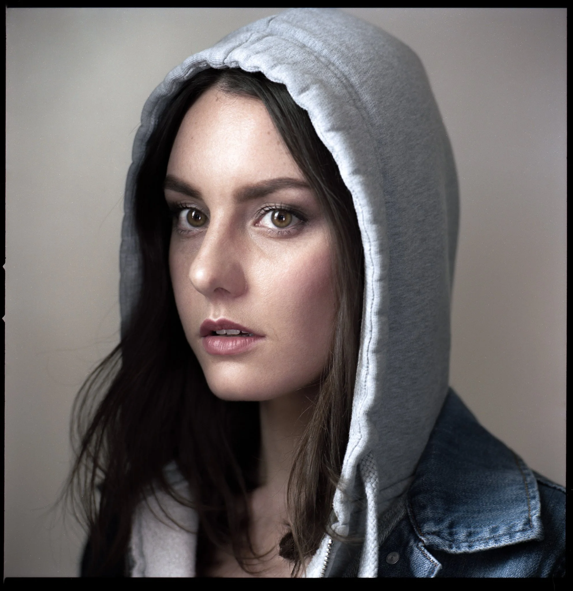 Close-up of a young woman with fair skin, wearing a gray hooded sweatshirt and a denim jacket, against a neutral background.