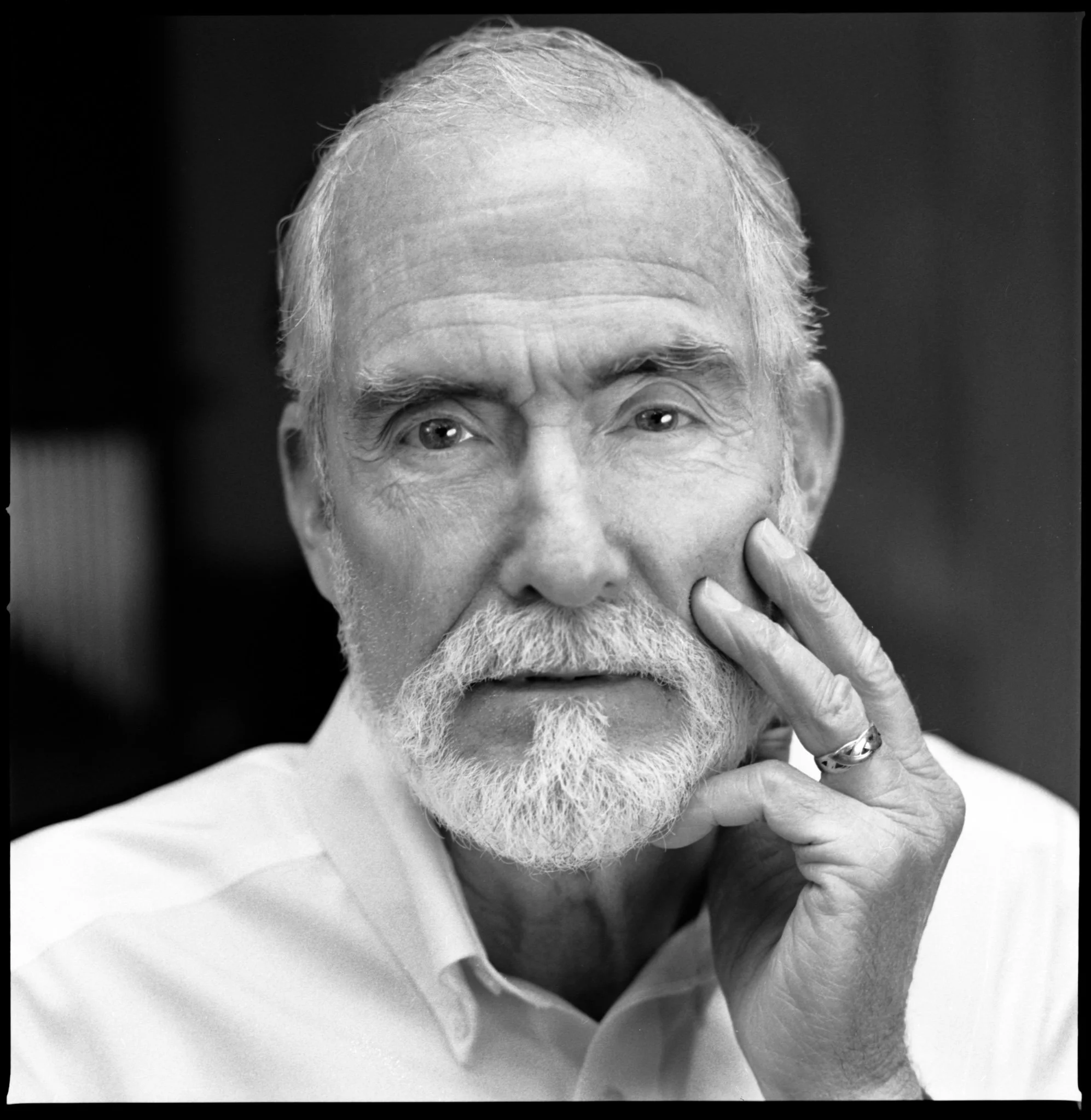 Black and white portrait of an elderly man with a beard, wearing a collared shirt, resting his face on his hand with fingers touching his cheek, looking directly at the camera.
