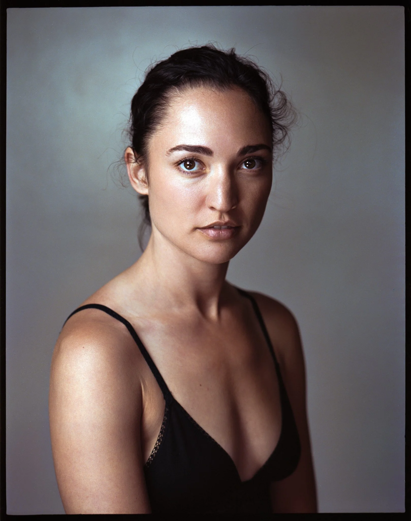 Portrait of a young woman with dark hair in a black spaghetti strap top, looking at the camera with a neutral expression.