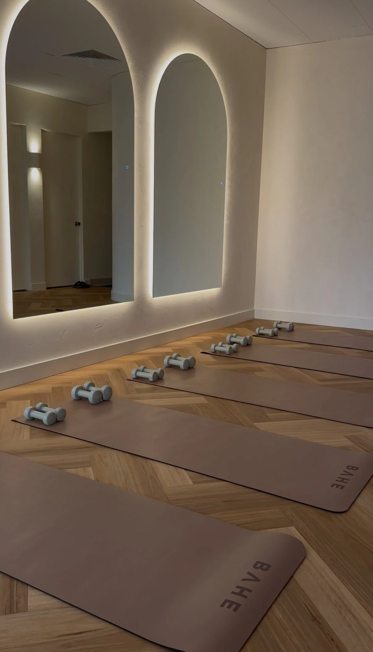 Yoga mats and small dumbbells arranged on a wooden floor in a modern, well-lit room with a large mirror and arched wall cutouts.