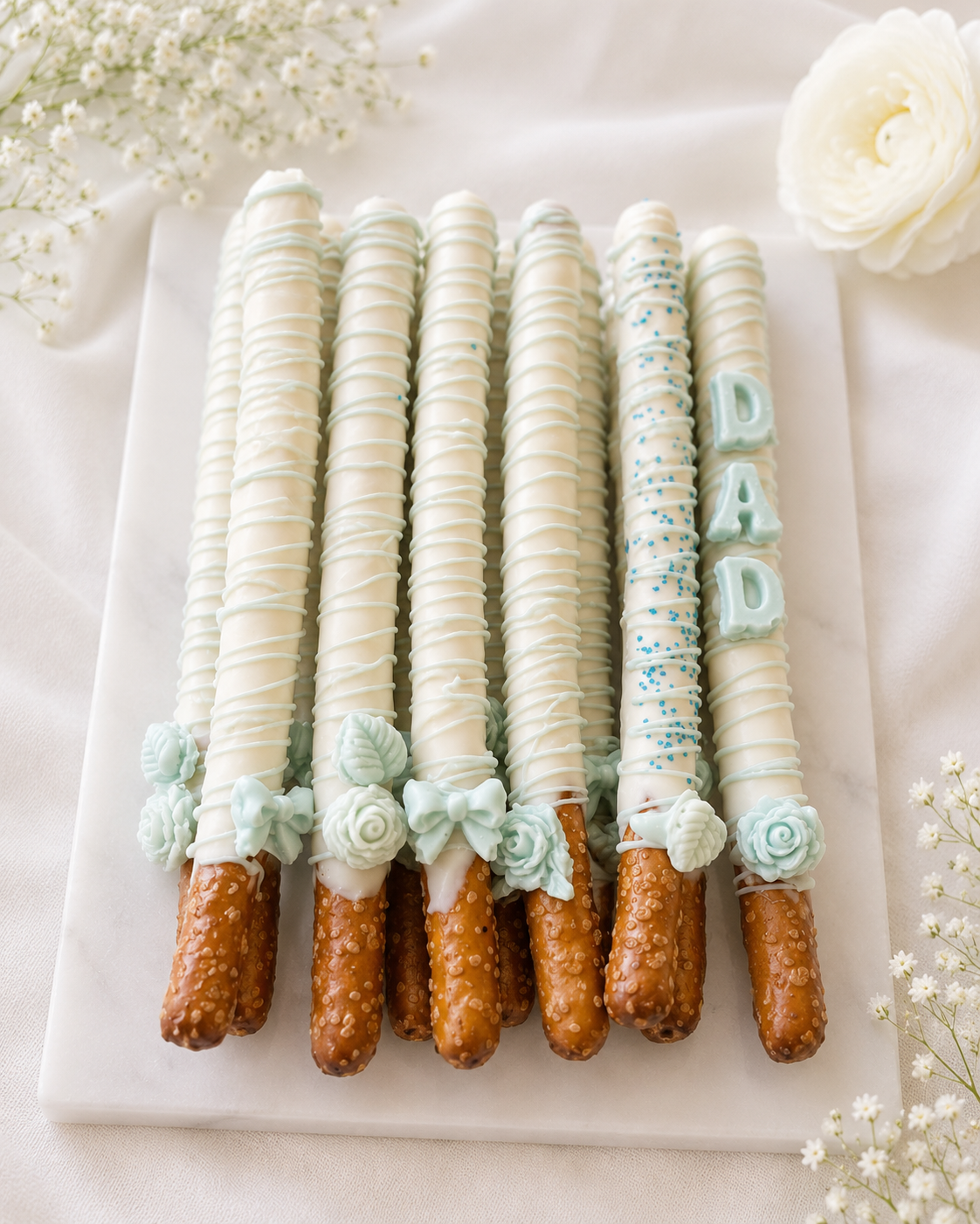 Decorated pretzel rods dipped in creamy white chocolate with soft blue drizzle, floral accents, bows, and “DAD” lettering, arranged on an elegant white platter.