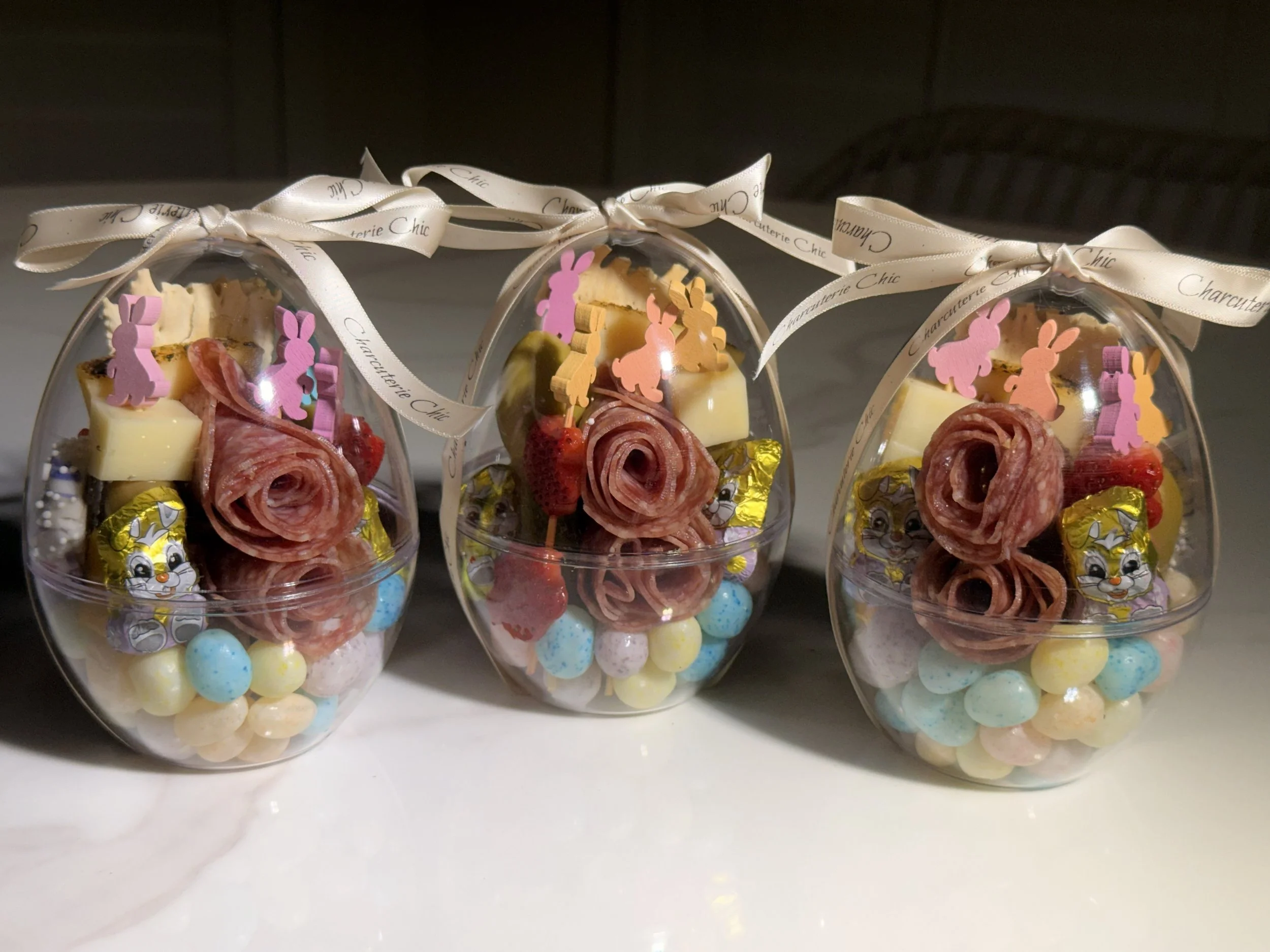 Easter-themed individual charcuterie egg containers with salami roses, cheeses, fruit, pastel candies and bunny details, finished with Charcuterie Chic ribbon.