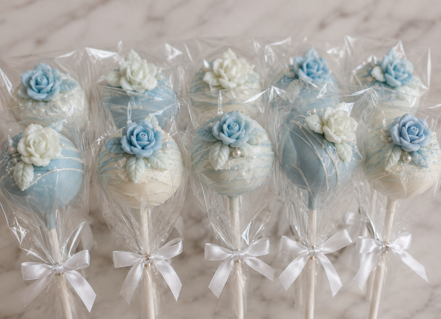 Round cake pops dipped in white and soft blue chocolate with marbled designs, delicate drizzle, sprinkles, and floral accents.