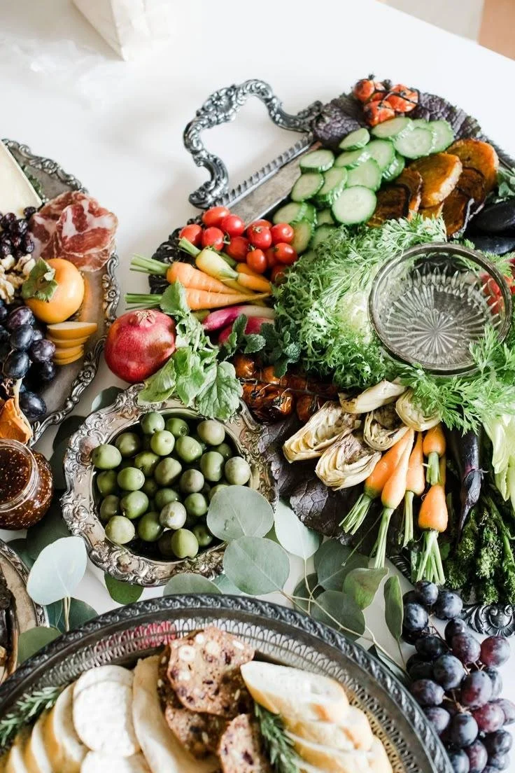 Elegant grazing table with fresh vegetables, olives, artisan meats, fruit and greenery displayed on vintage silver serving pieces.