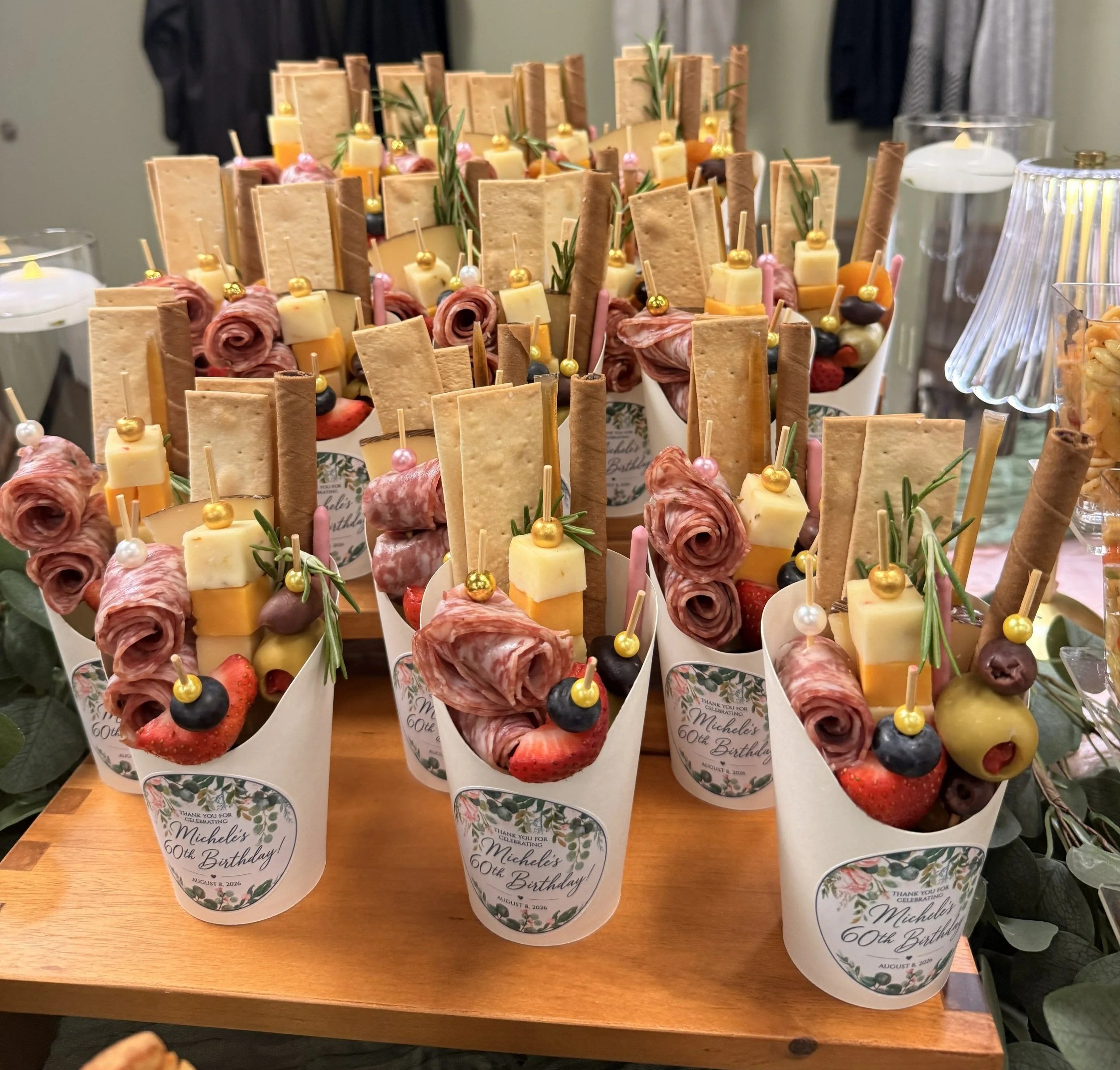 Custom birthday charcuterie cups displayed on wooden risers with salami roses, artisan cheeses, crackers, fruit and personalized labels.