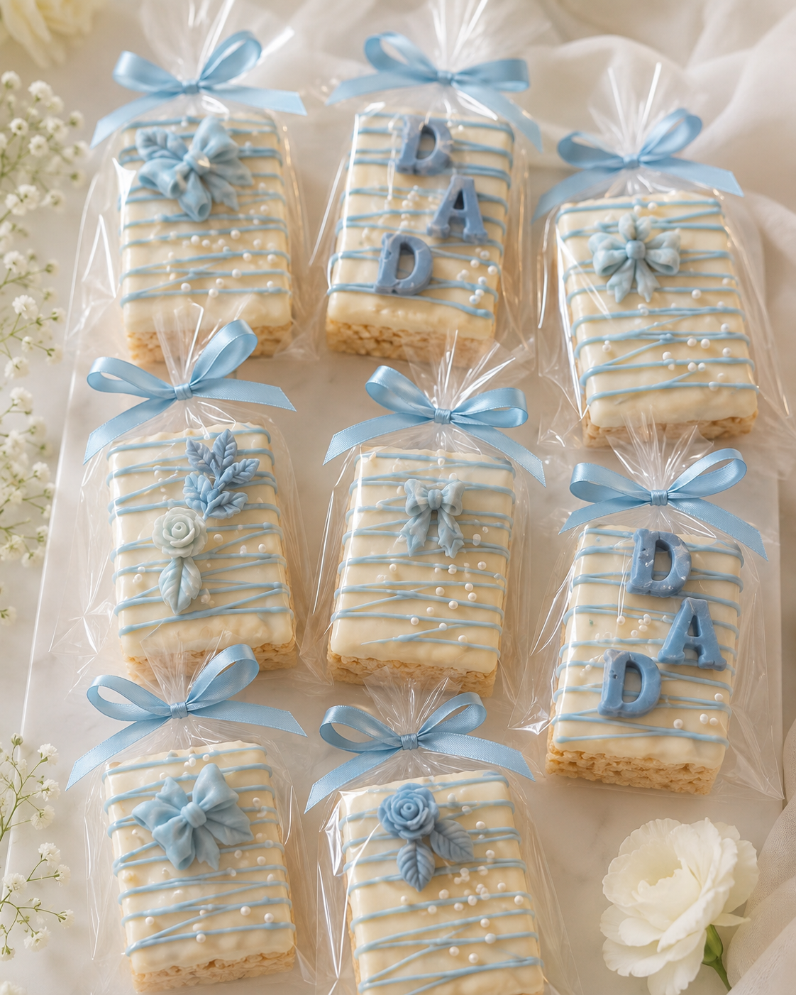 White chocolate-covered Rice Krispie treats decorated with soft blue drizzle, pearl sprinkles, floral accents, bows, and “DAD” lettering, individually wrapped with blue ribbons.