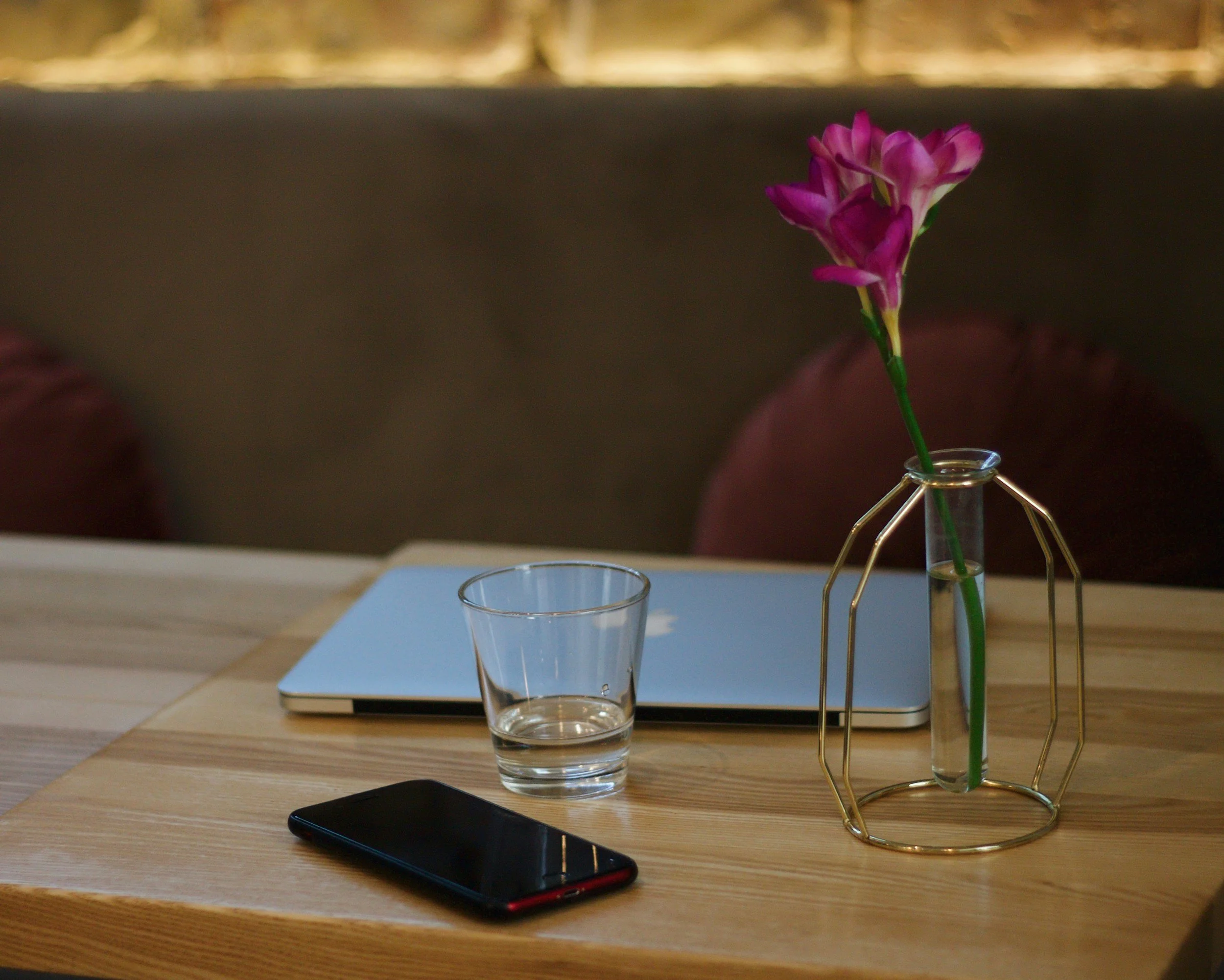 A wooden table with a closed silver laptop, a clear glass of water, a black smartphone, and a unique geometric glass vase holding pink flowers.