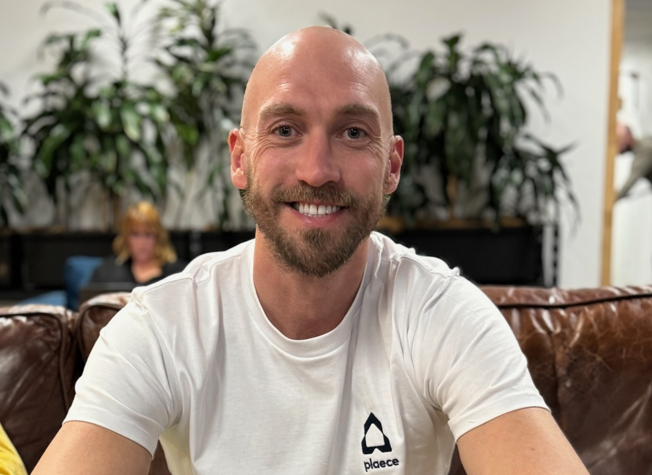 Founder Paul Elderkin, a smiling man with a bald head and beard, sitting on a brown leather couch, wearing a white t-shirt with a logo, with greenery and a woman in the background.