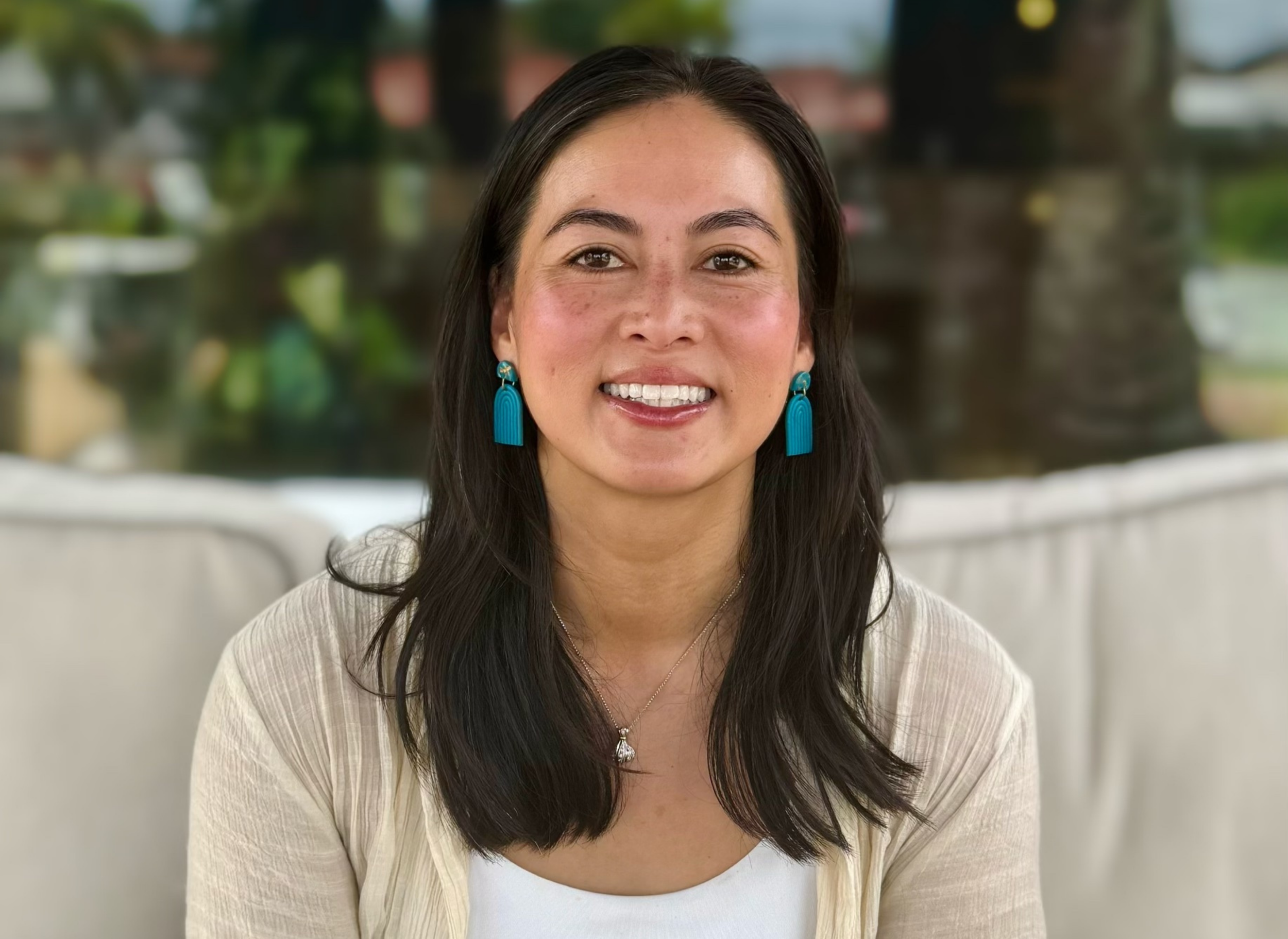Founder Ann Luong, a smiling woman with dark hair wearing blue earrings and a light cream cardigan, sitting indoors with a blurred outdoor background.