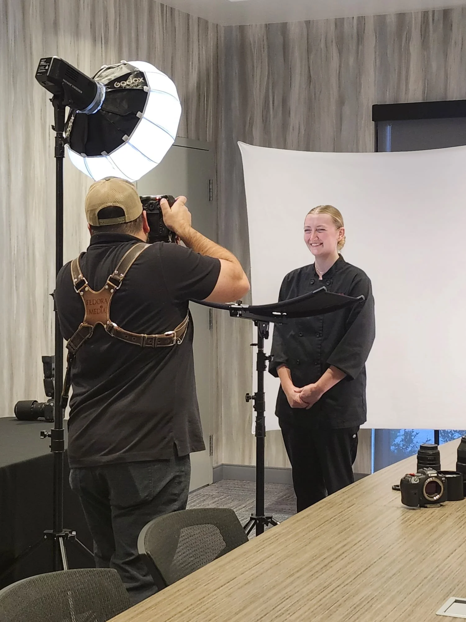 Photographer taking a picture of a smiling woman in a professional photoshoot setting with studio lights and a white backdrop.