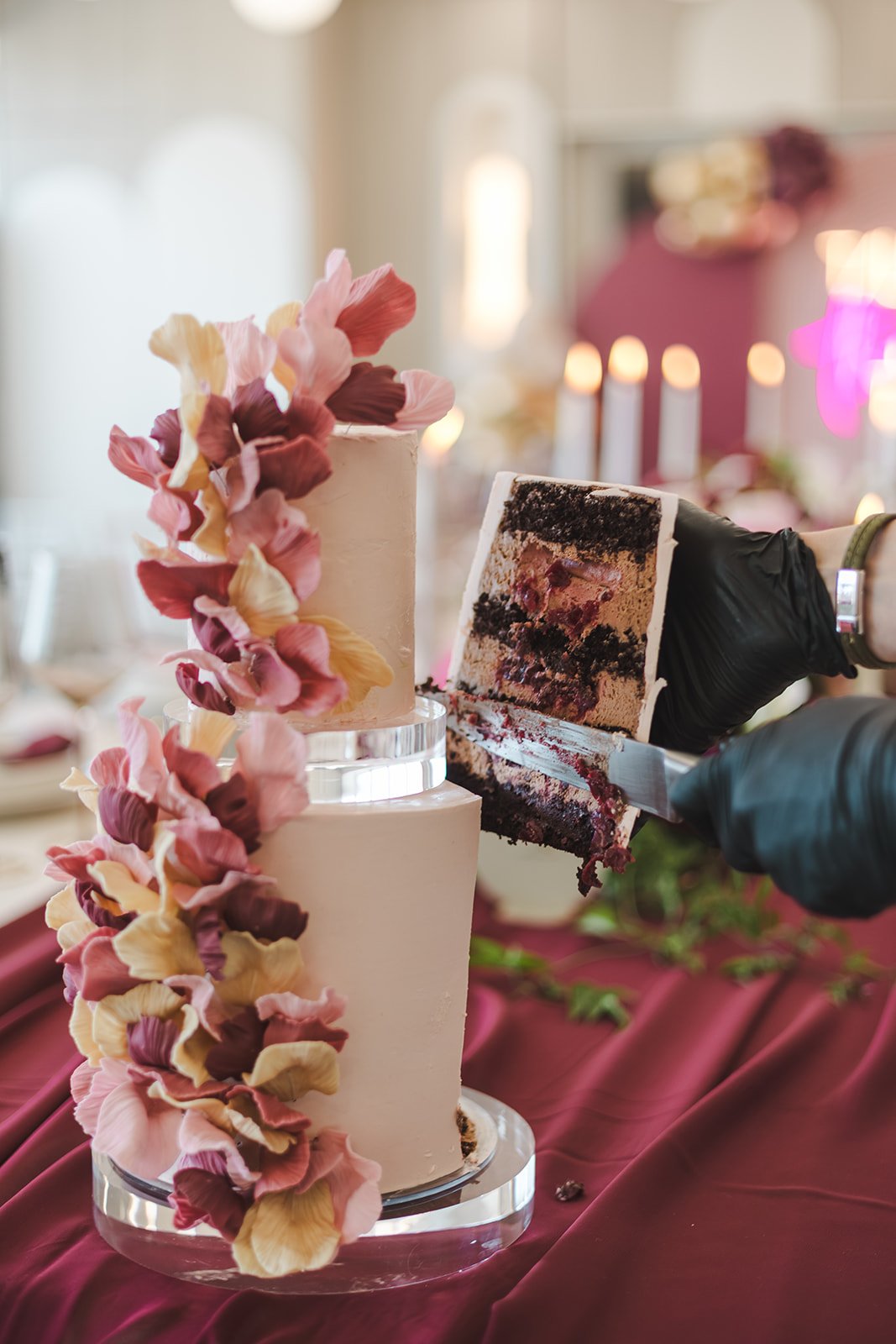 A person in black gloves is cutting a slice from a multi-layered cake decorated with pink, purple, and cream-colored edible flowers. The cake has smooth white frosting and is set on a reflective cake stand on a burgundy tablecloth.