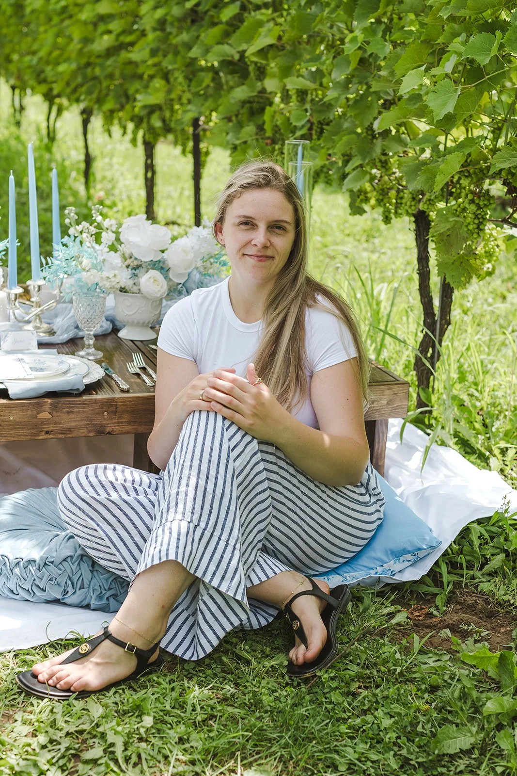 A woman sitting on the grass near a decorated outdoor table with white flowers, candles, and blue accents, surrounded by lush green grapevines.