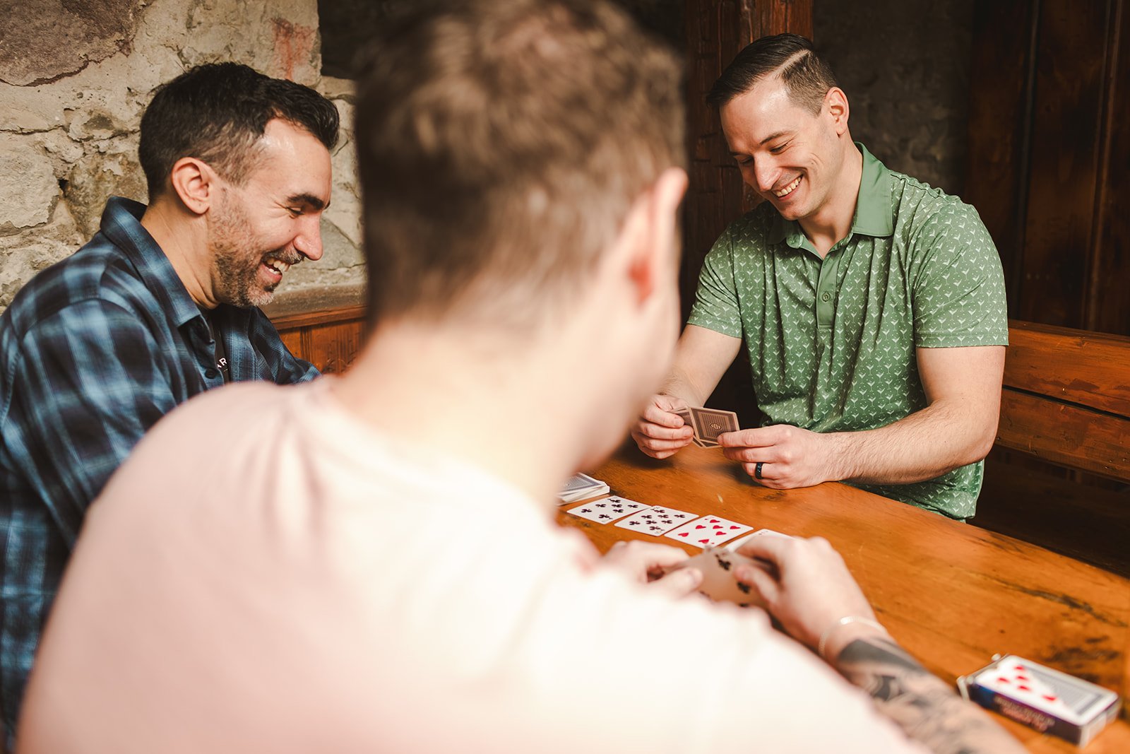 Four people playing cards and laughing around a wooden table in a cozy, rustic room.