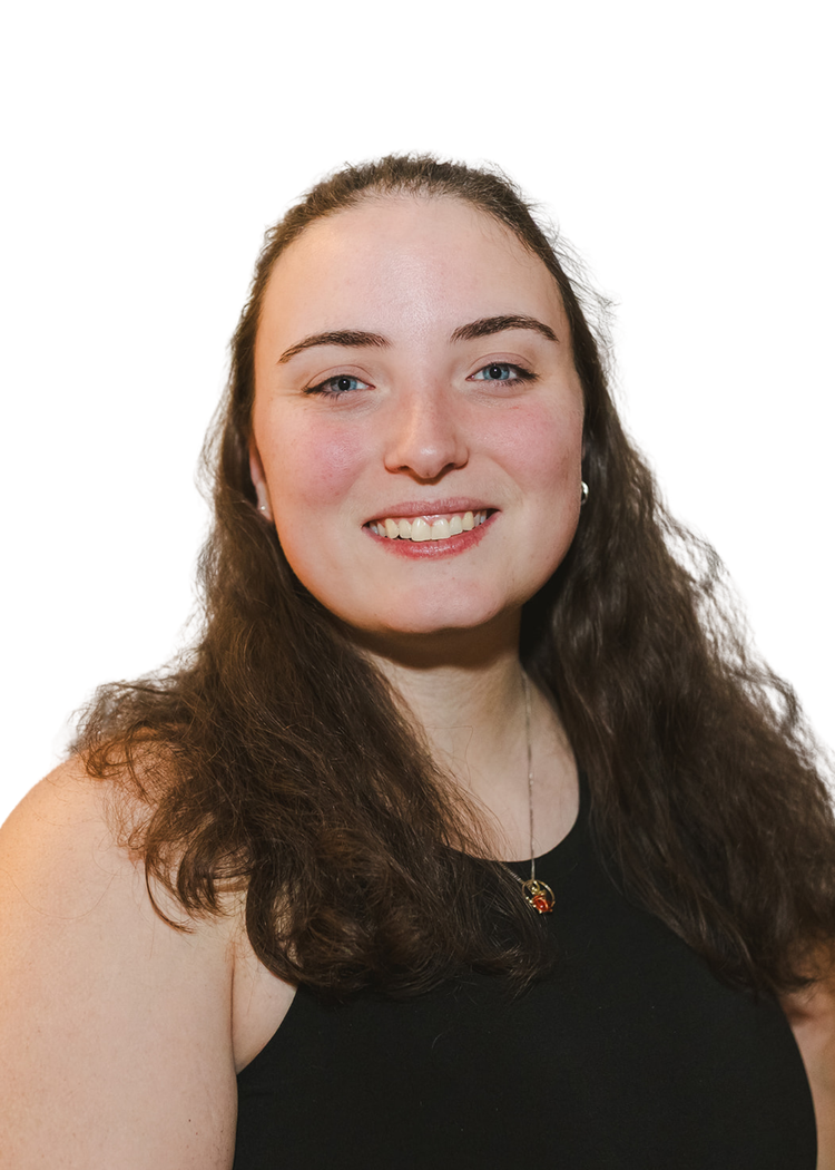 A woman with long, wavy brown hair wearing a black sleeveless top and a necklace with a small pendant, smiling against a plain white background.