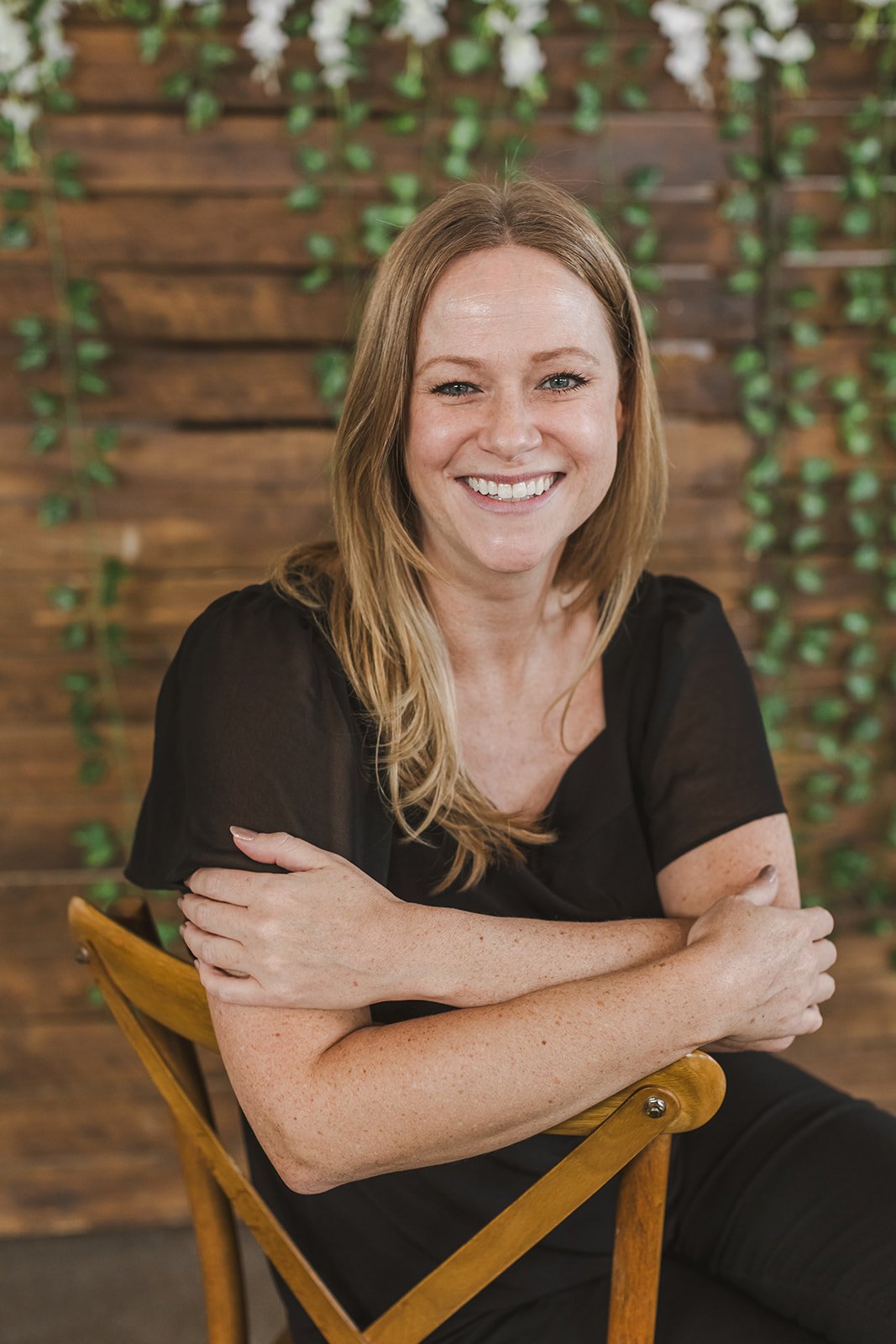 A smiling woman with long red hair sitting on a wooden chair with arms crossed, against a background of wooden panels with green vines.