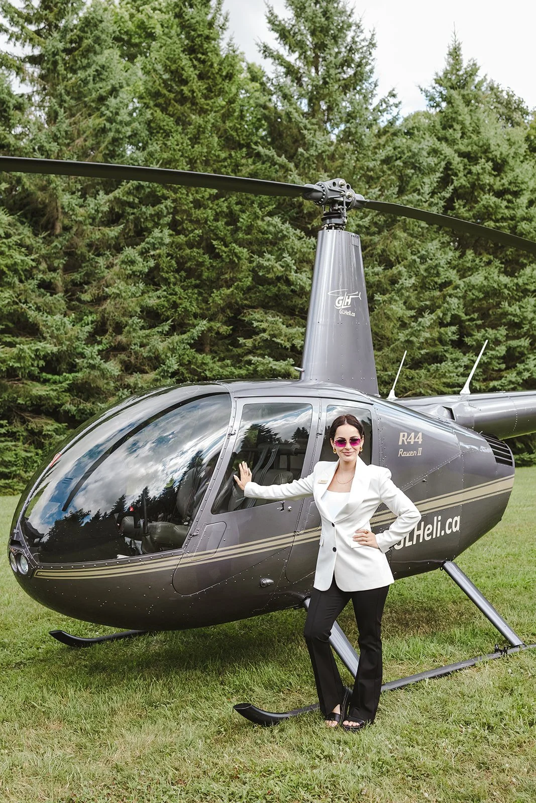 A woman in a white blazer, black pants, and pink sunglasses standing next to a gray helicopter on a grassy field with trees in the background.