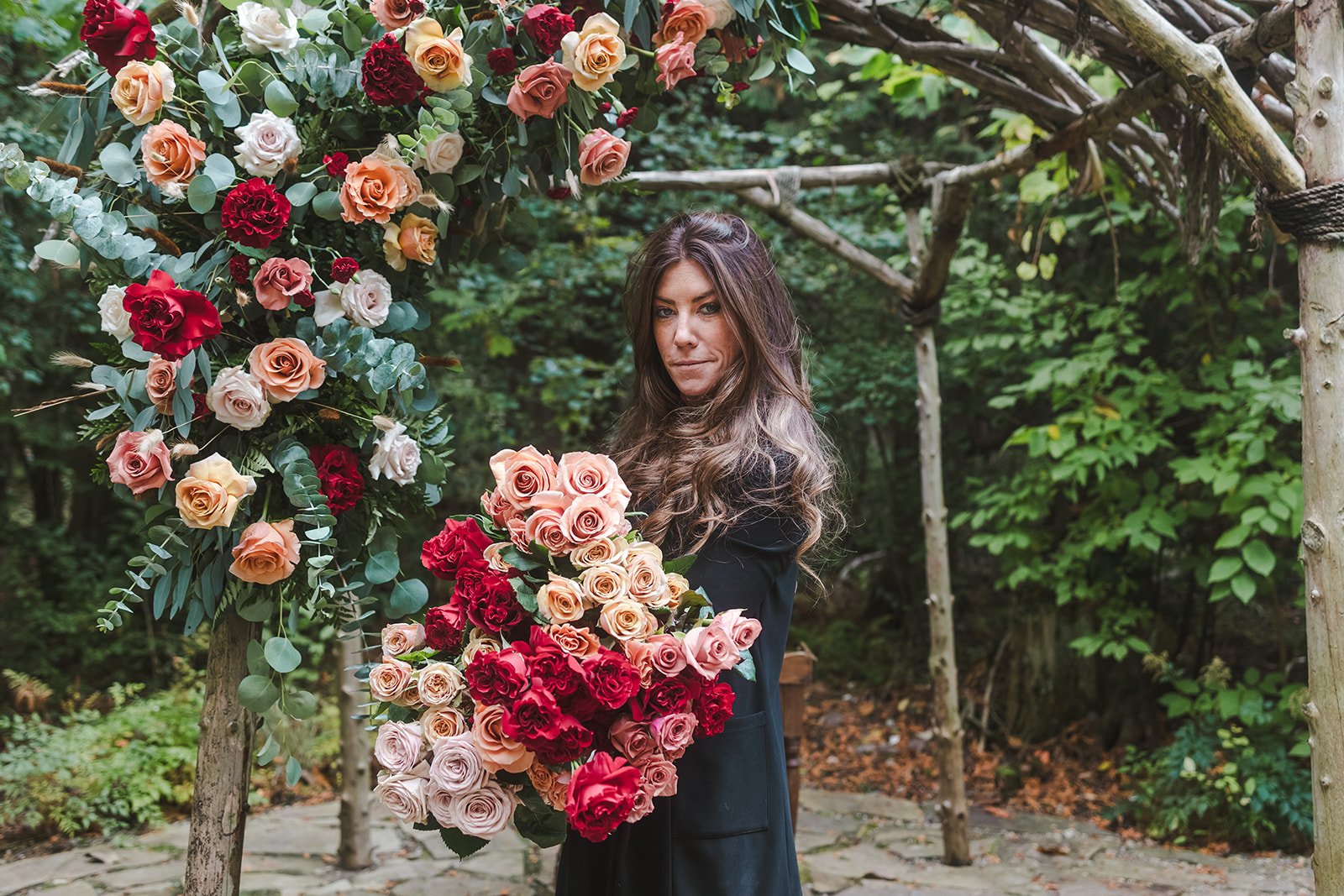 A woman with long wavy brown hair standing outdoors amidst a floral display of peach, pink, white, and red roses. She is dressed in a black coat and looking directly at the camera with a neutral expression, surrounded by green foliage and a wooden arch decorated with flowers.