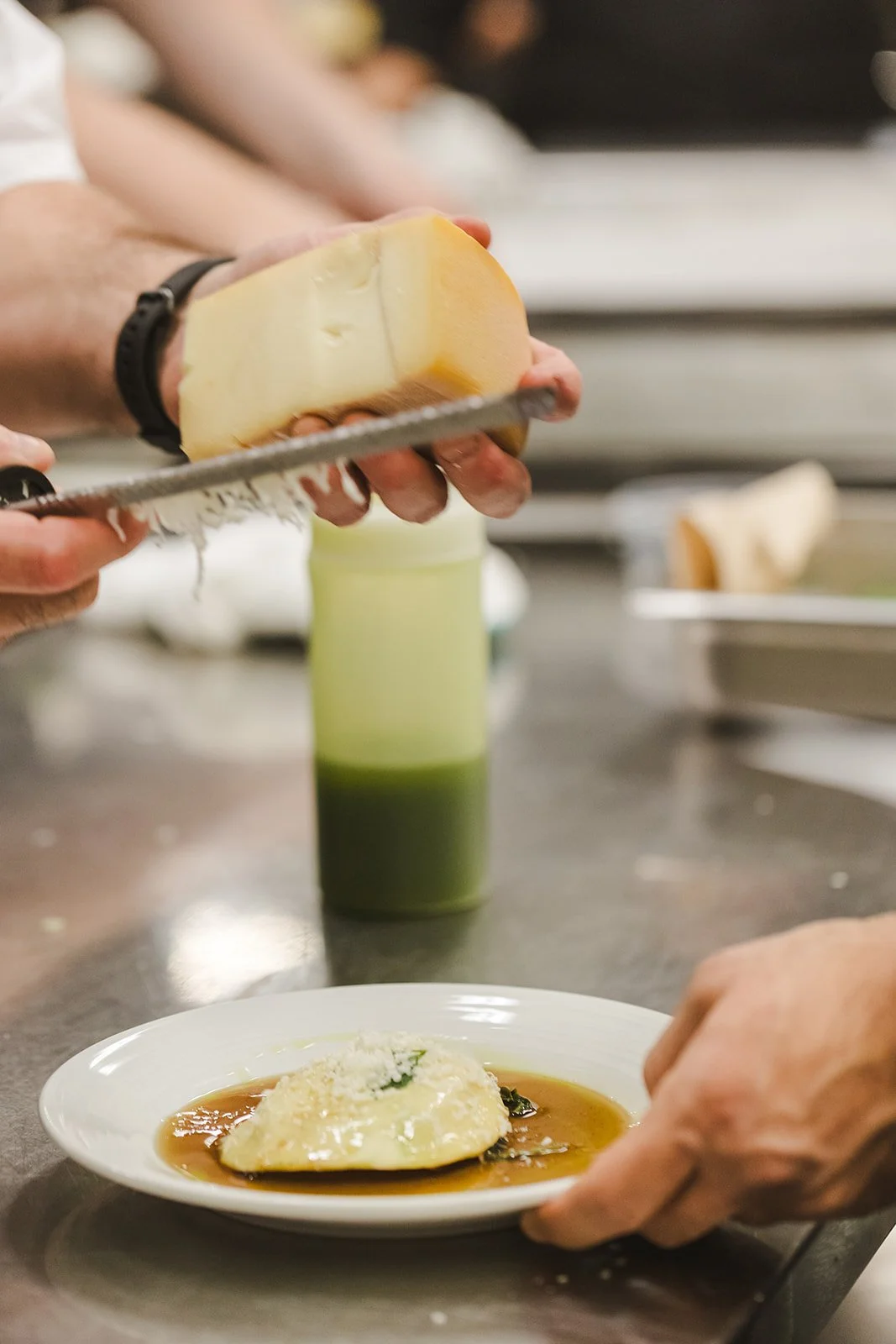 Person grating cheese over a plate of ravioli with sauce, with green juice in the background.