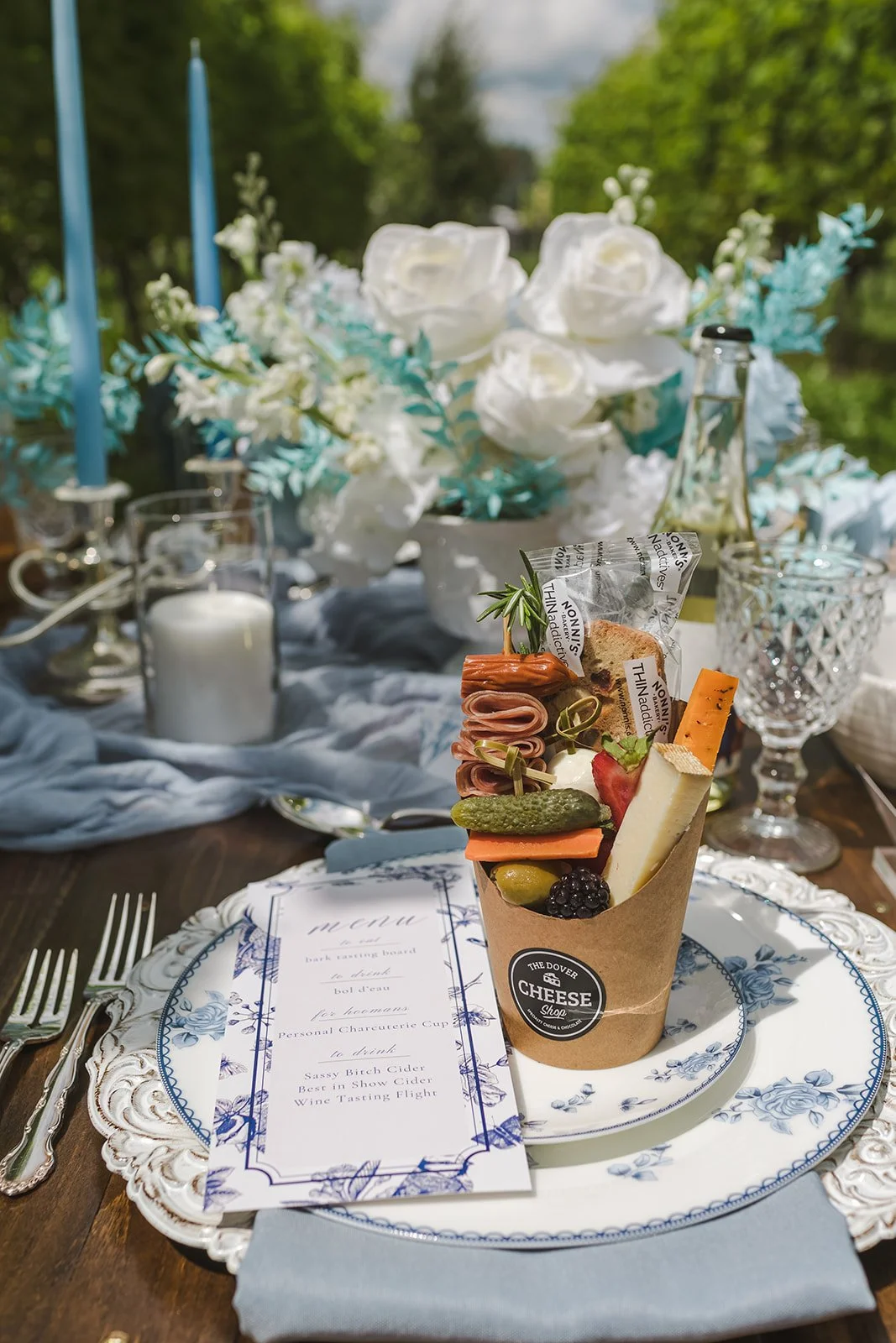 A food tower resembling a cheese board on a fancy table with white and blue floral dishware, a menu, and elegant glassware in an outdoor setting with trees and a cloudy sky