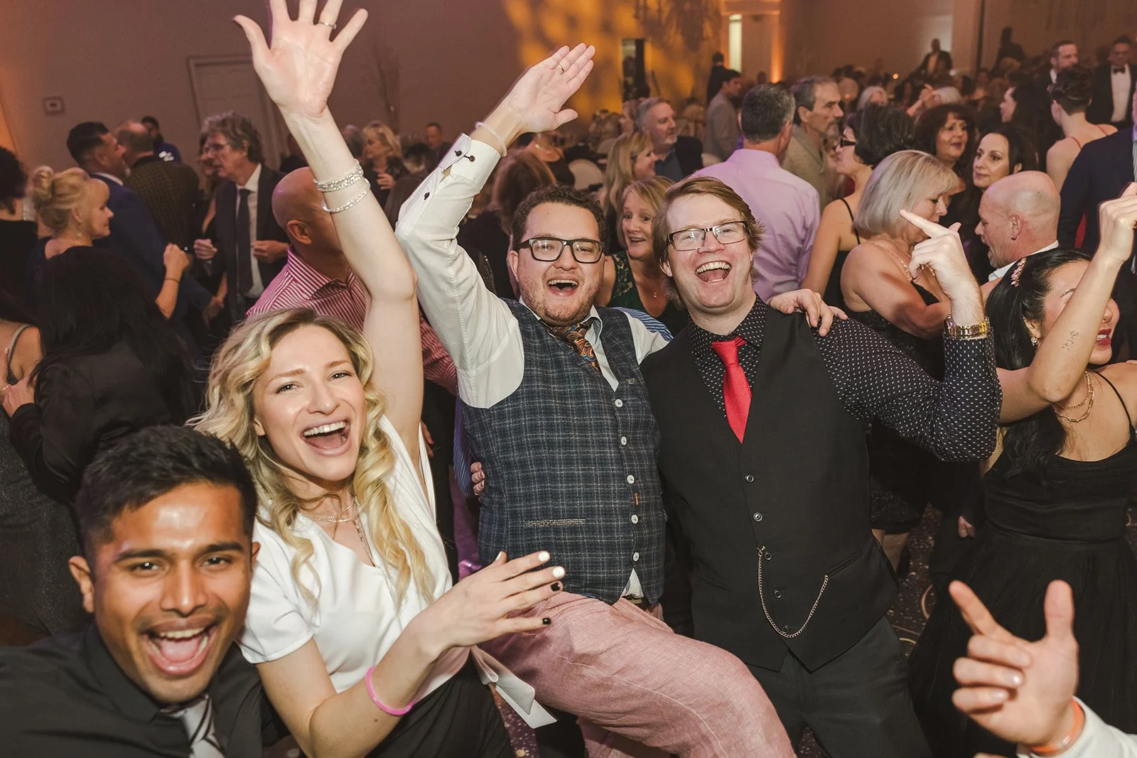 Group of people enjoying a lively event or party, smiling, laughing, and dancing in a crowded indoor venue with warm lighting.