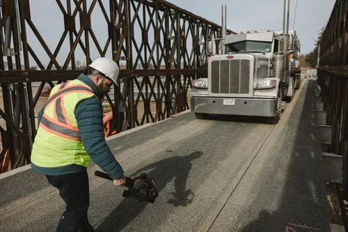Construction worker using a handheld tool on a bridge with trucks passing by.