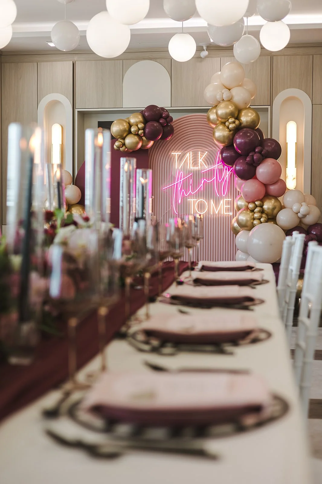 Decorated event space with a neon sign that reads 'Talk Thirty to Me' and a balloon arch in gold, pink, and purple hues. A long table set with plates, napkins, and glasses is in the foreground.