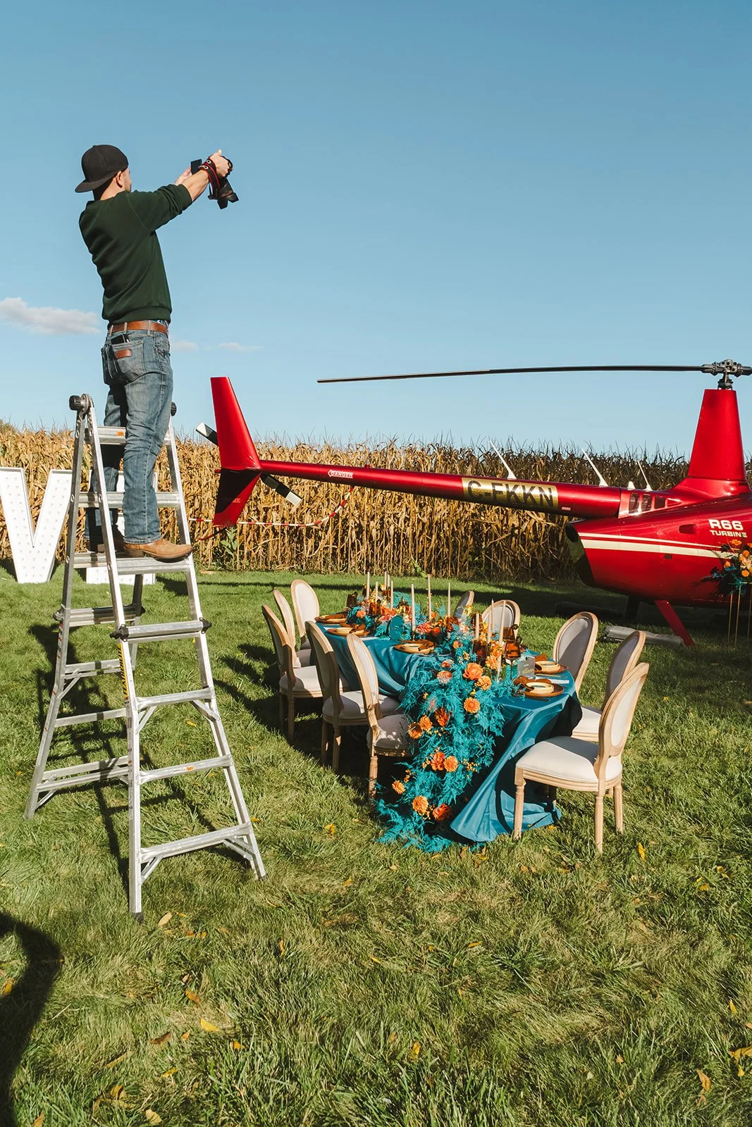 Person standing on ladder taking photo of a red helicopter at outdoor celebration with decorated table and chairs.