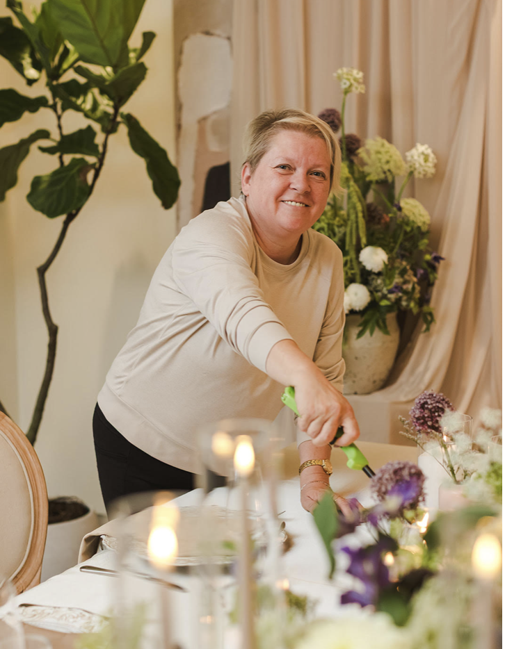 Woman smiling while cutting a floral centerpiece on a dining table.
