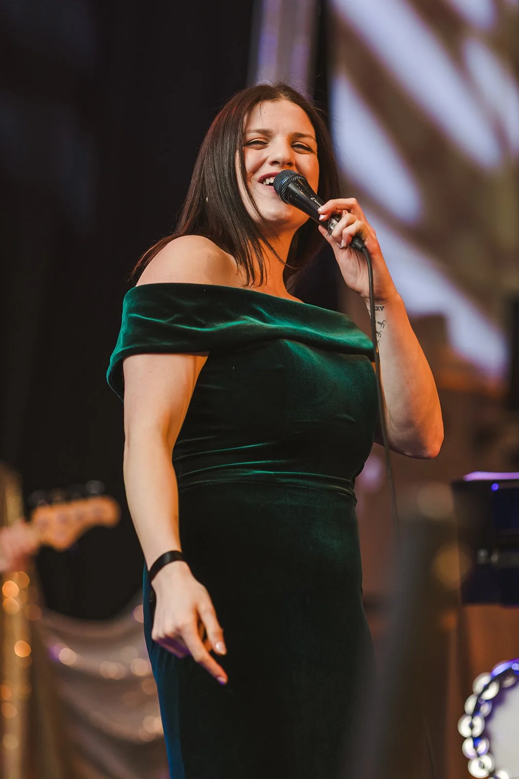 A woman in a dark green off-the-shoulder dress singing into a microphone on stage with a blurred background.
