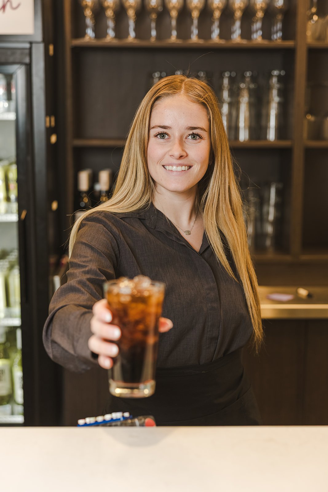 A smiling waitress in a black uniform offers a glass of soda with ice at a restaurant or bar counter.