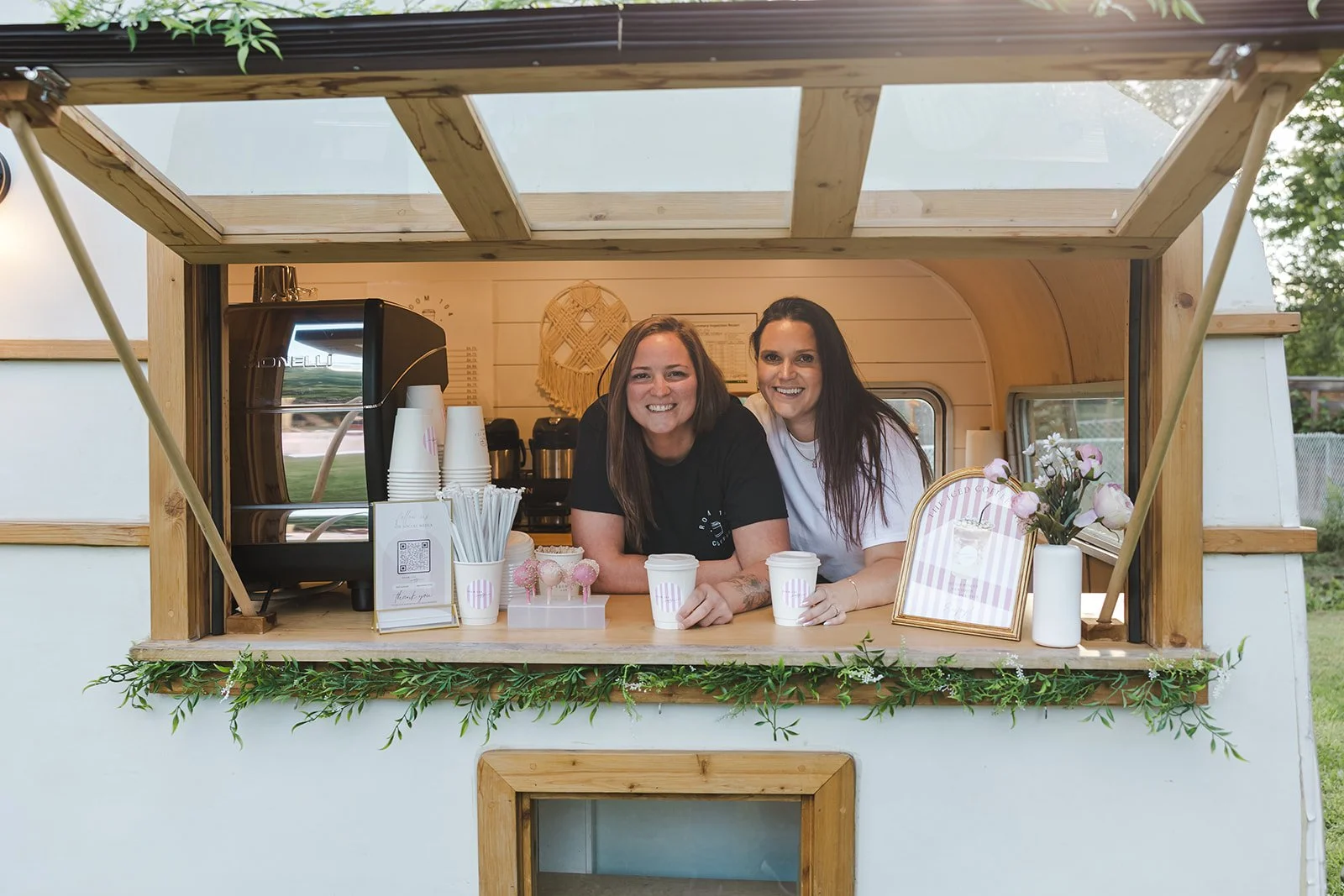 Two women standing behind a coffee stand smiling at the camera, with coffee cups and coffee machine inside.