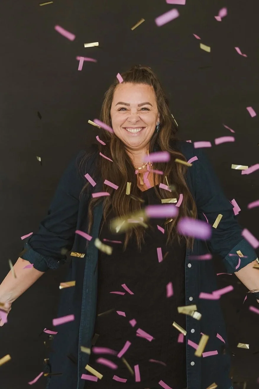A woman celebrating with pink and gold confetti falling around her, smiling joyfully against a black background.
