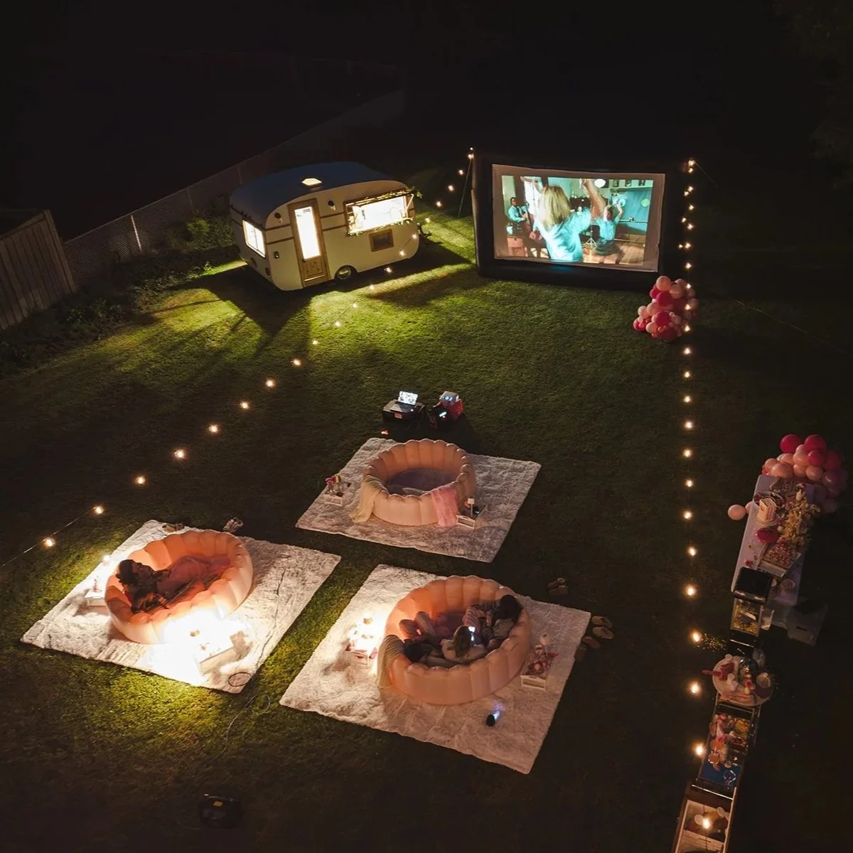 Night outdoor birthday party setup with illuminated string lights, a screen projecting a video, pink inflatable seats, a small caravan, and a snack table with balloons.