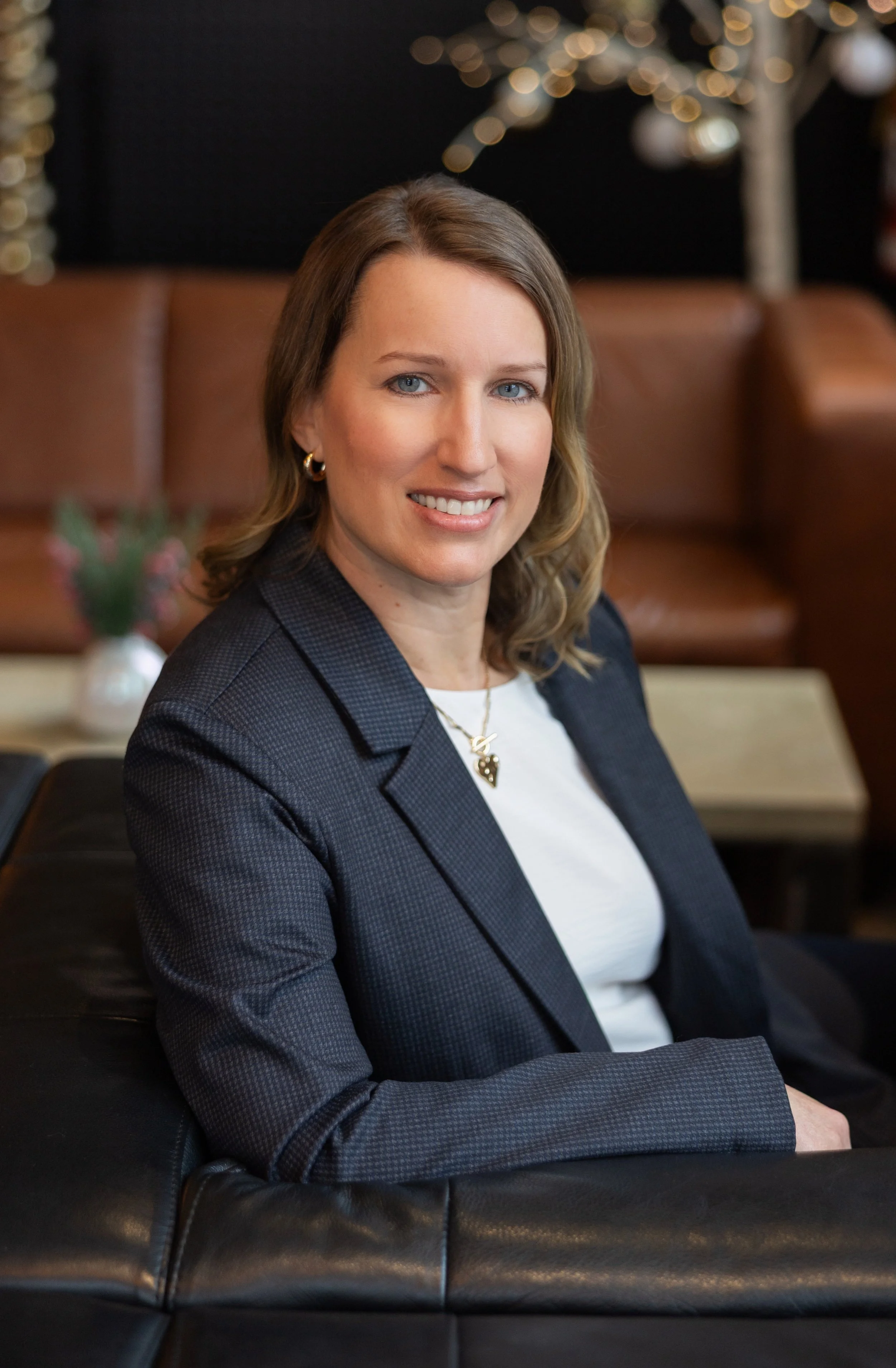 A woman with shoulder-length light brown hair, wearing a dark blazer and white shirt, sitting on a black leather couch, smiling at the camera with a blurred background of a decorated Christmas tree and lights.