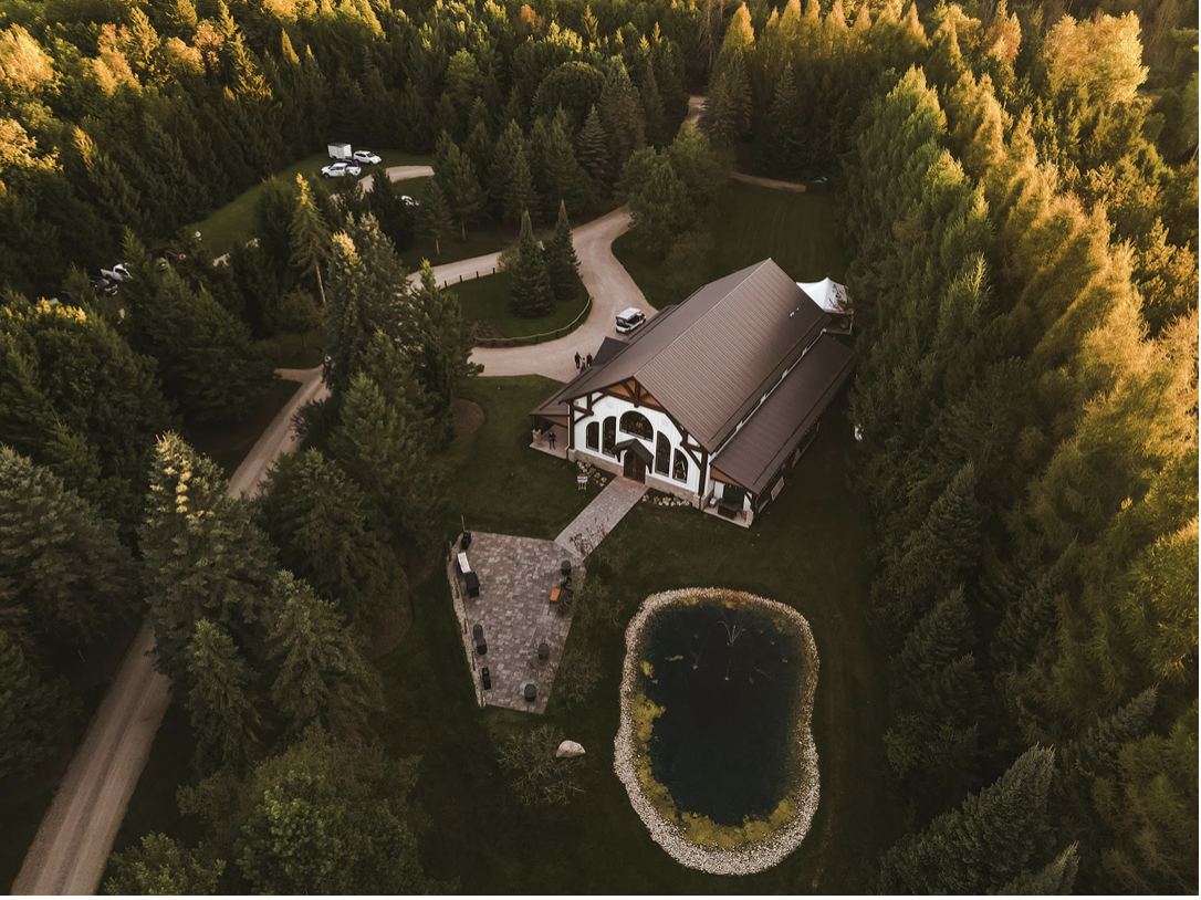 An aerial view of a house surrounded by dense trees, with a brick pathway leading to the front door and a small pond with a fountain in the yard. There are a few parked cars near the house and additional buildings or structures nearby.