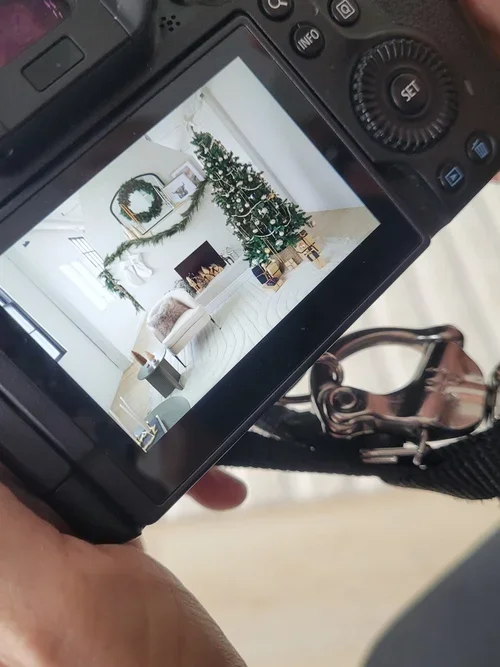 A camera's LCD screen showing a decorated Christmas tree in a living room with white walls, a fireplace, and holiday decorations.