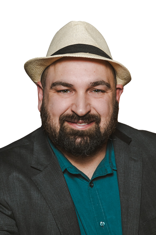 A smiling man with a beard and mustache wearing a beige fedora hat, a dark blazer, and a teal shirt, posing against a white background.