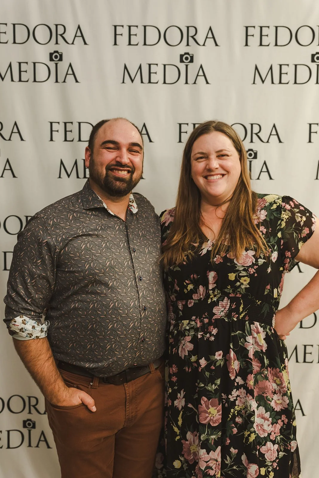 A man and woman smiling and standing close together in front of a backdrop with the text 'FEDORA MEDIA' repeated multiple times.