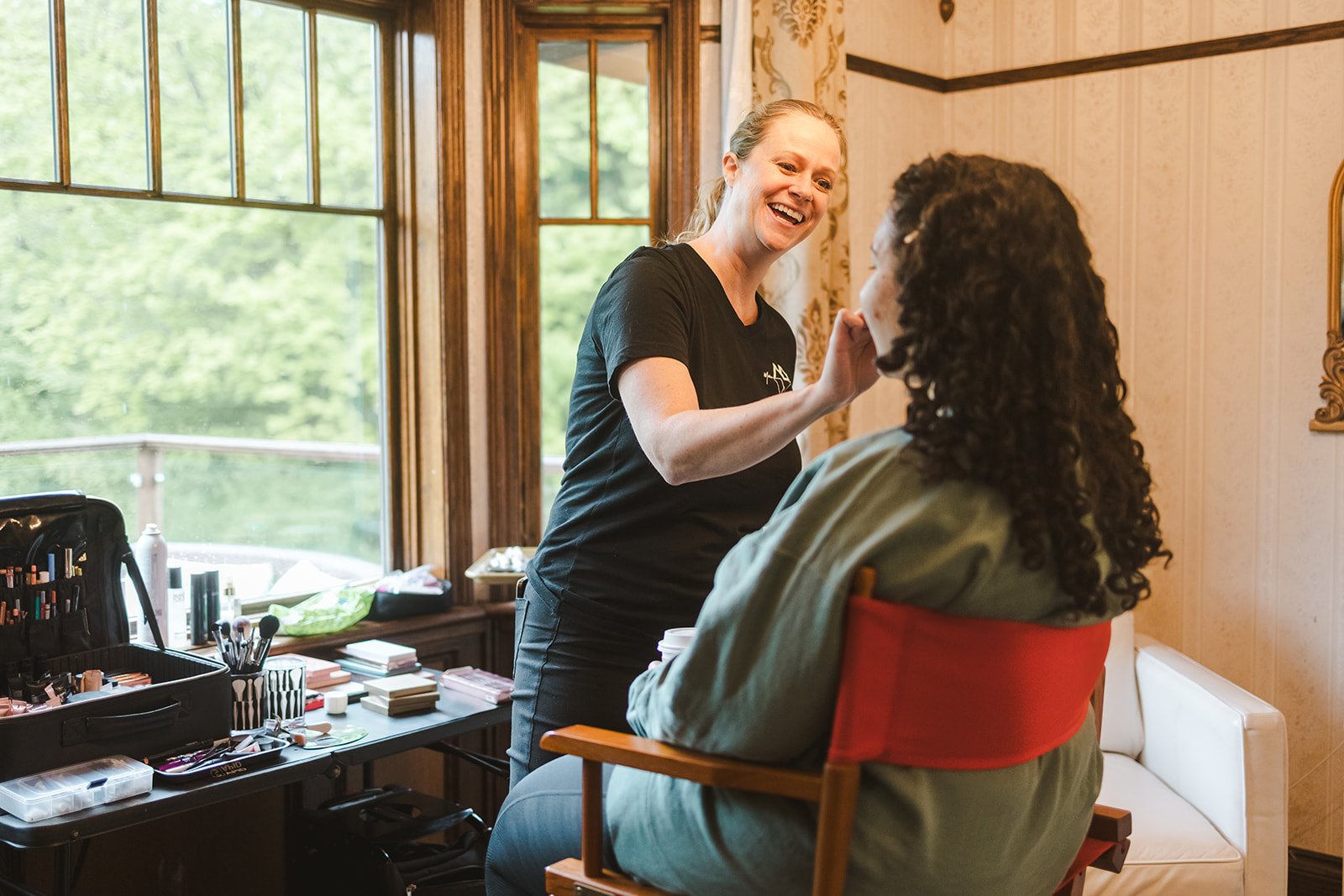 Makeup artist applying makeup to a seated woman in a room with wooden window frames and patterned wallpaper, outdoors visible through the window.