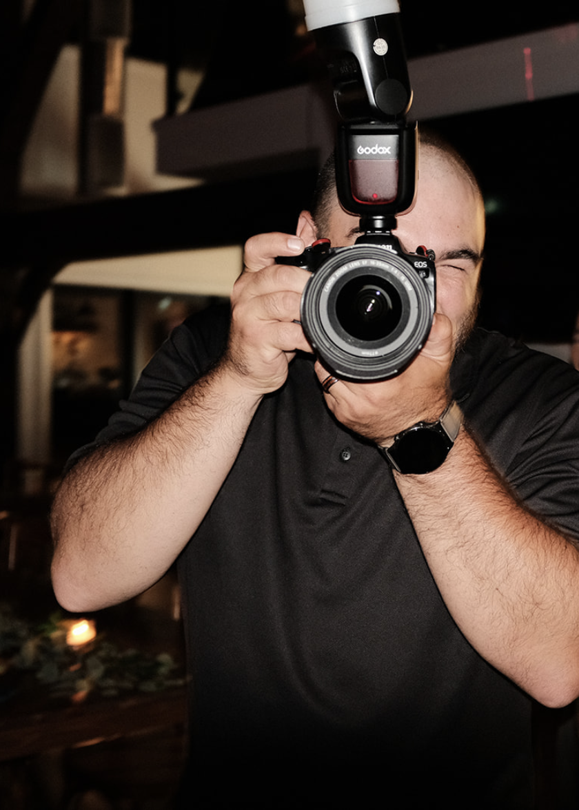 Photographer taking a picture indoors, holding a professional camera with a flash attachment, wearing a black shirt and a smartwatch.