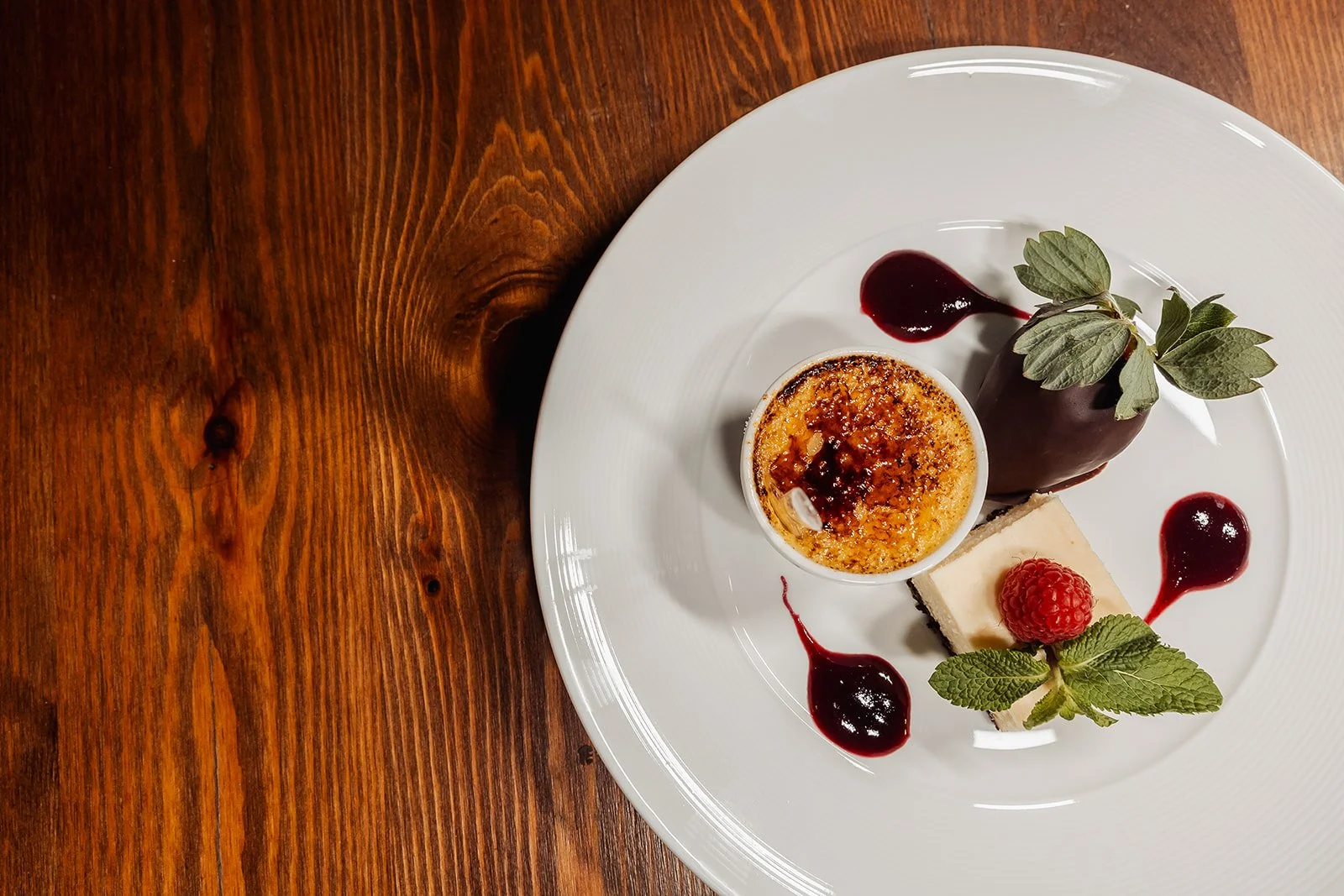 Dessert plate featuring crème brûlée, a chocolate-covered cake with mint leaves, and a cheesecake with a raspberry and mint garnish, all with berry sauce drizzles on a white plate.
