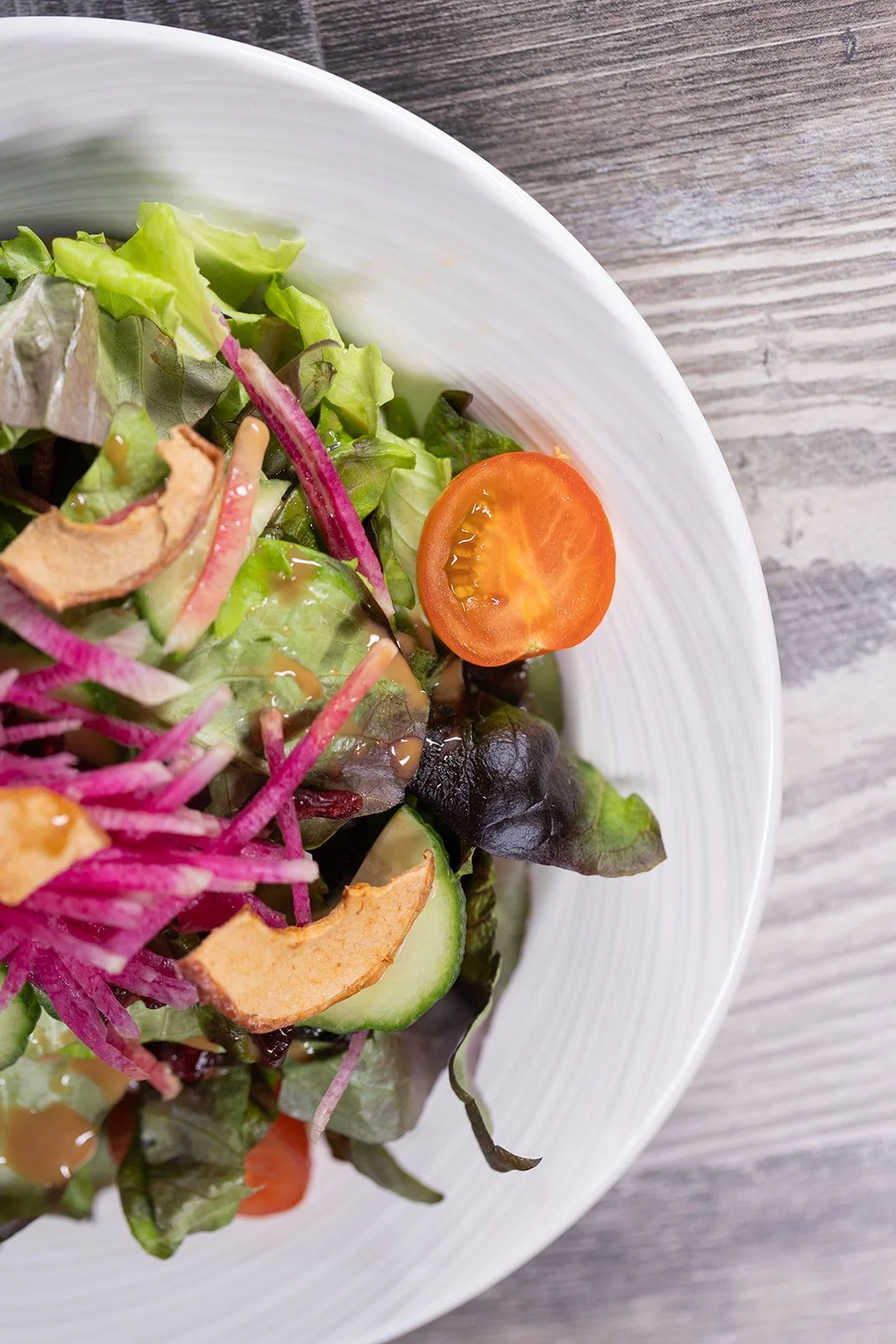 Fresh salad with cherry tomato, cucumbers, mixed greens, shredded purple cabbage, and sliced apples in a white bowl on a wooden surface.