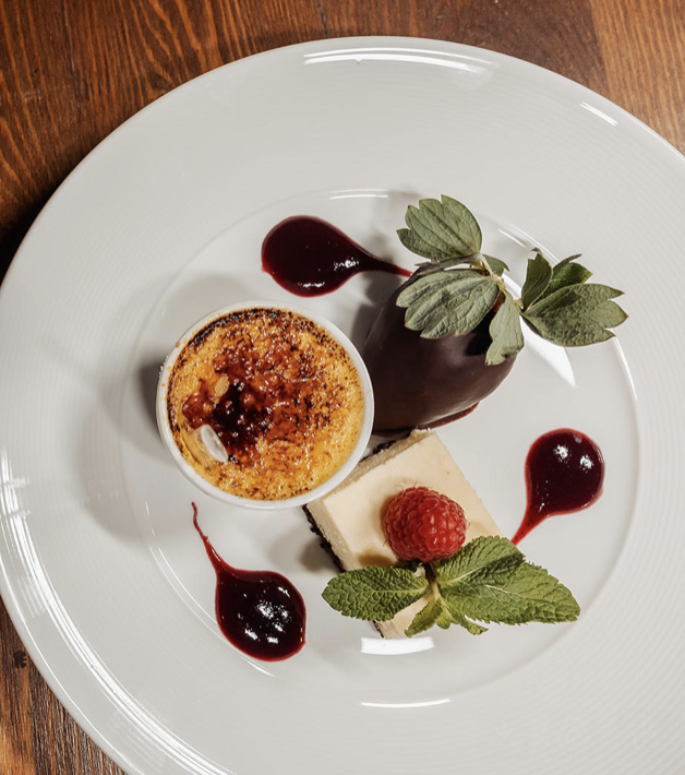 Dessert plate with a chocolate-covered pastry topped with a sprig of mint, a slice of cheesecake garnished with a raspberry and mint leaves, and small dollops of raspberry sauce on a white plate