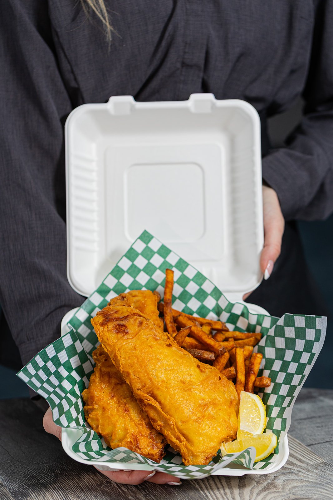 A takeout container with fried fish, French fries, and lemon wedges, held by a person in a dark shirt.