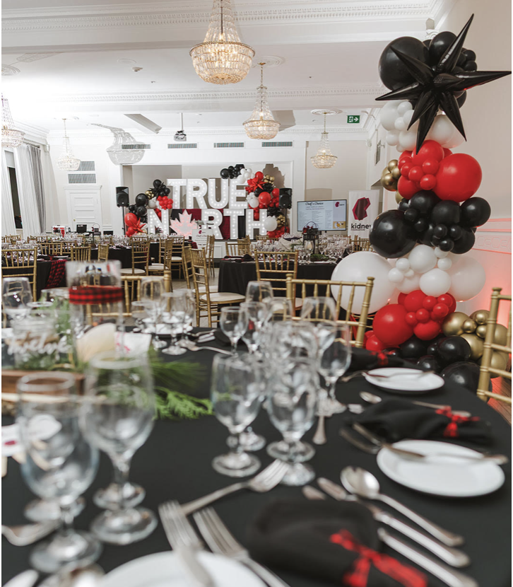 A banquet hall decorated with black, red, white, and gold balloons, and a large sign reading "True North" with a maple leaf, indicating a Canadian theme or celebration, set with neatly arranged tables, glassware, and gold chairs.