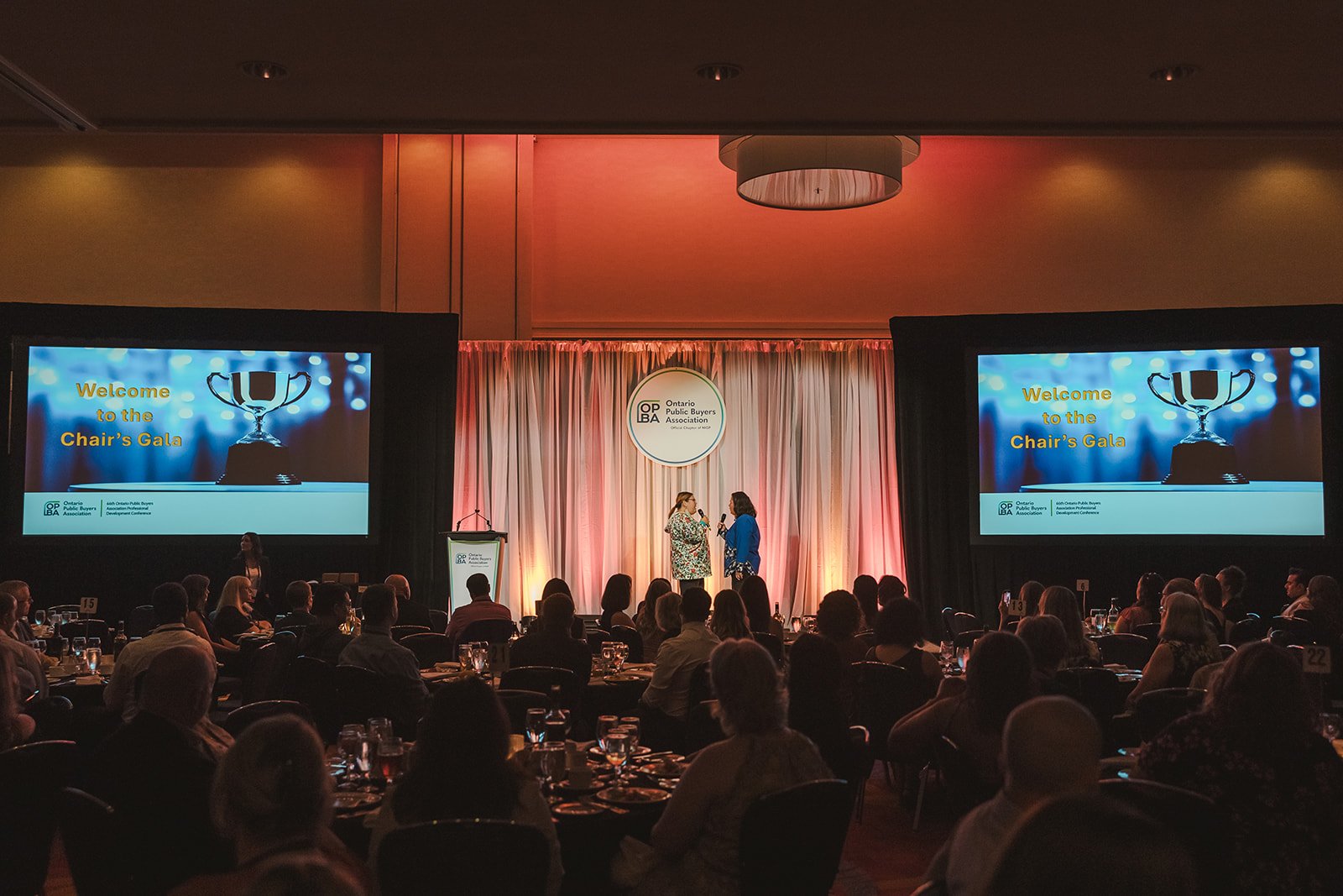 Audience seated at round tables facing a stage with two women speaking into microphones; large screens display 'Welcome to the Chair's Gala' and a trophy logo; central curtain backdrop with 'Ontario Public Buyers Association' logo.